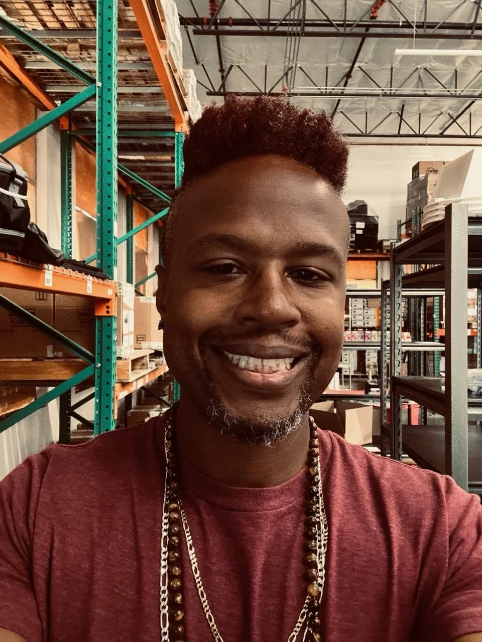 A smiling man with a beard and short curly hair, wearing a maroon t-shirt and layered necklaces, posing inside a warehouse with metal shelving and boxes.