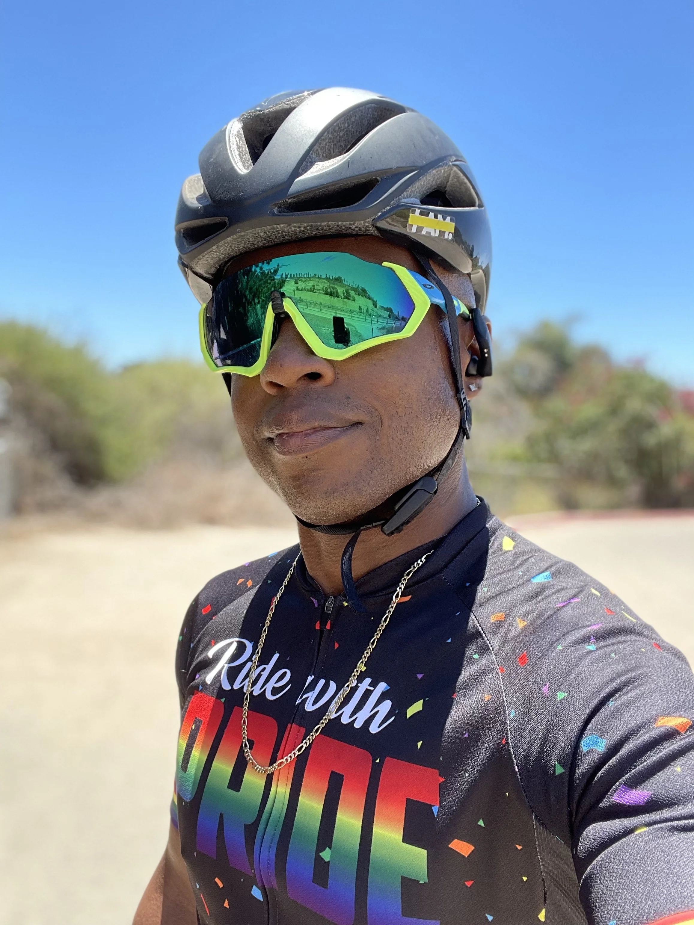 A person wearing a cycling helmet and colorful sunglasses takes a selfie outdoors, with a clear blue sky and some blurred greenery in the background.