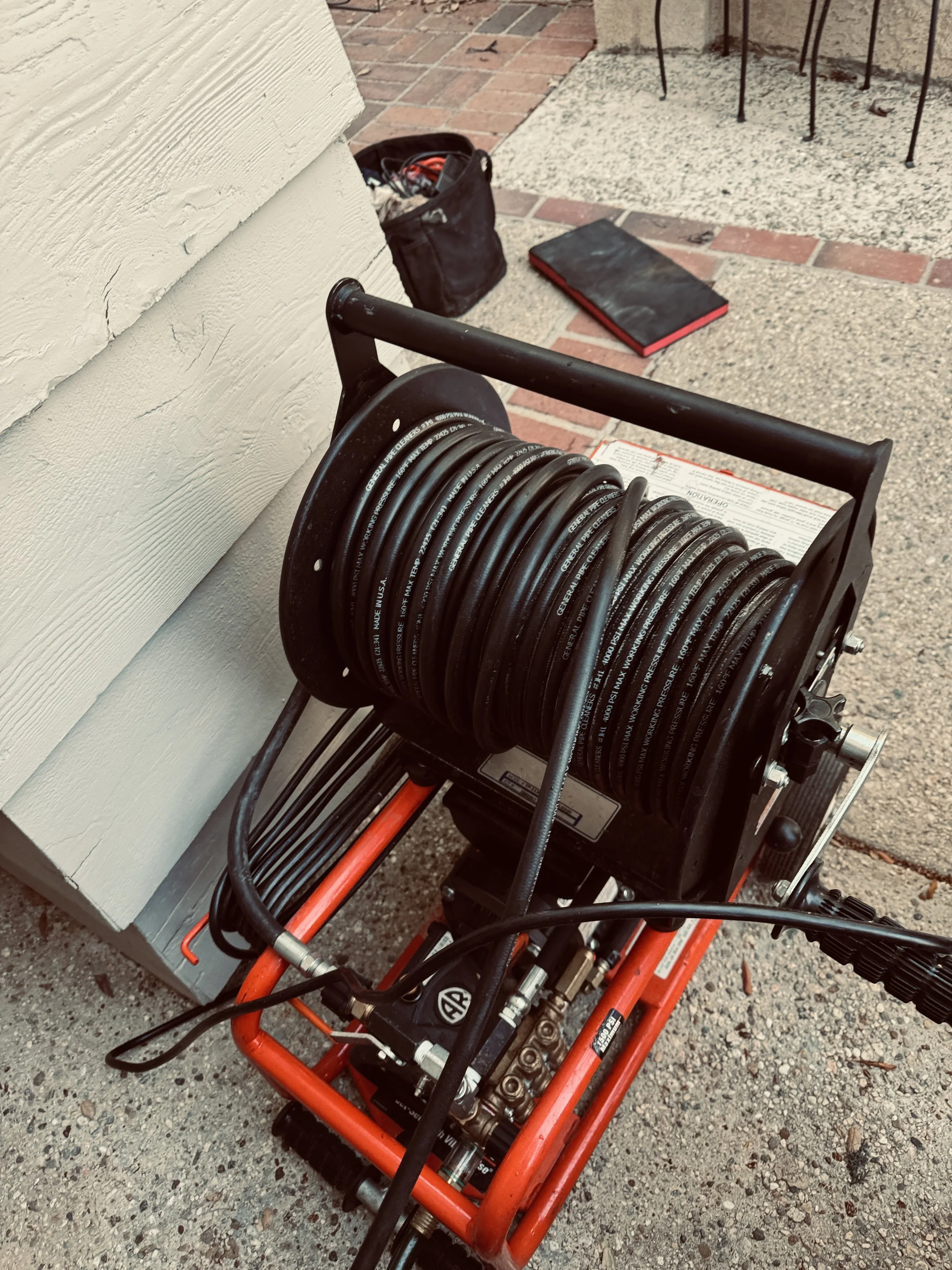 A pressure washer with a hose reel, red frame, and attached hydraulic equipment, positioned on a gravel surface outside near a building wall and a brick patio.