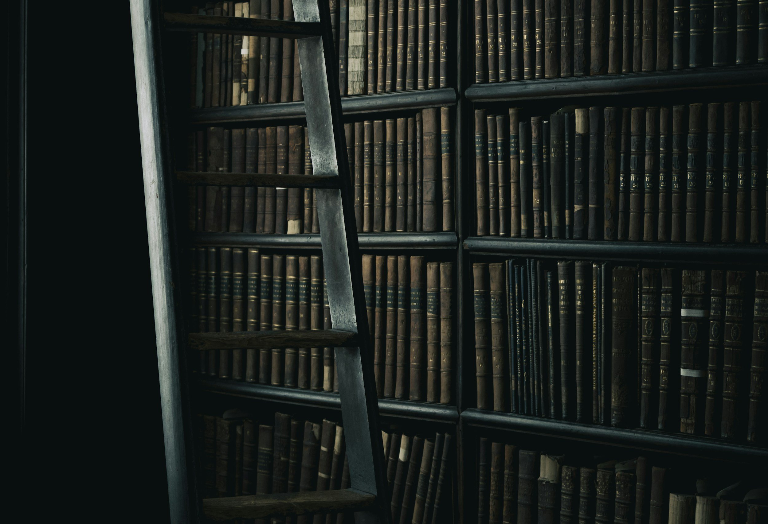 A bookshelf filled with old, leather-bound books and a wooden ladder leaning against it.