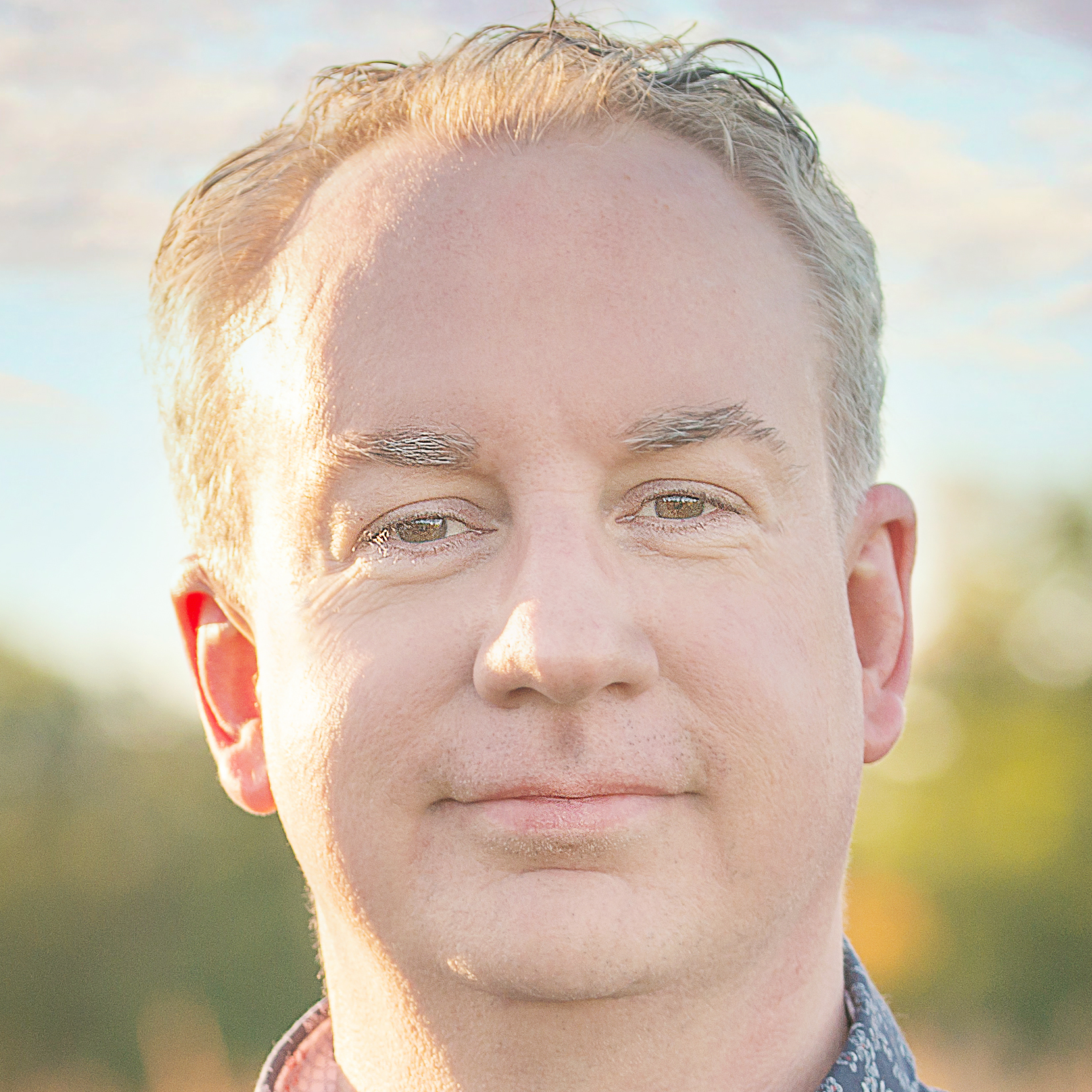 Close-up of Nick Richbell's face outdoors with sunlight, showing light-colored hair, blue eyes, and a slight smile, with a blurred nature background.