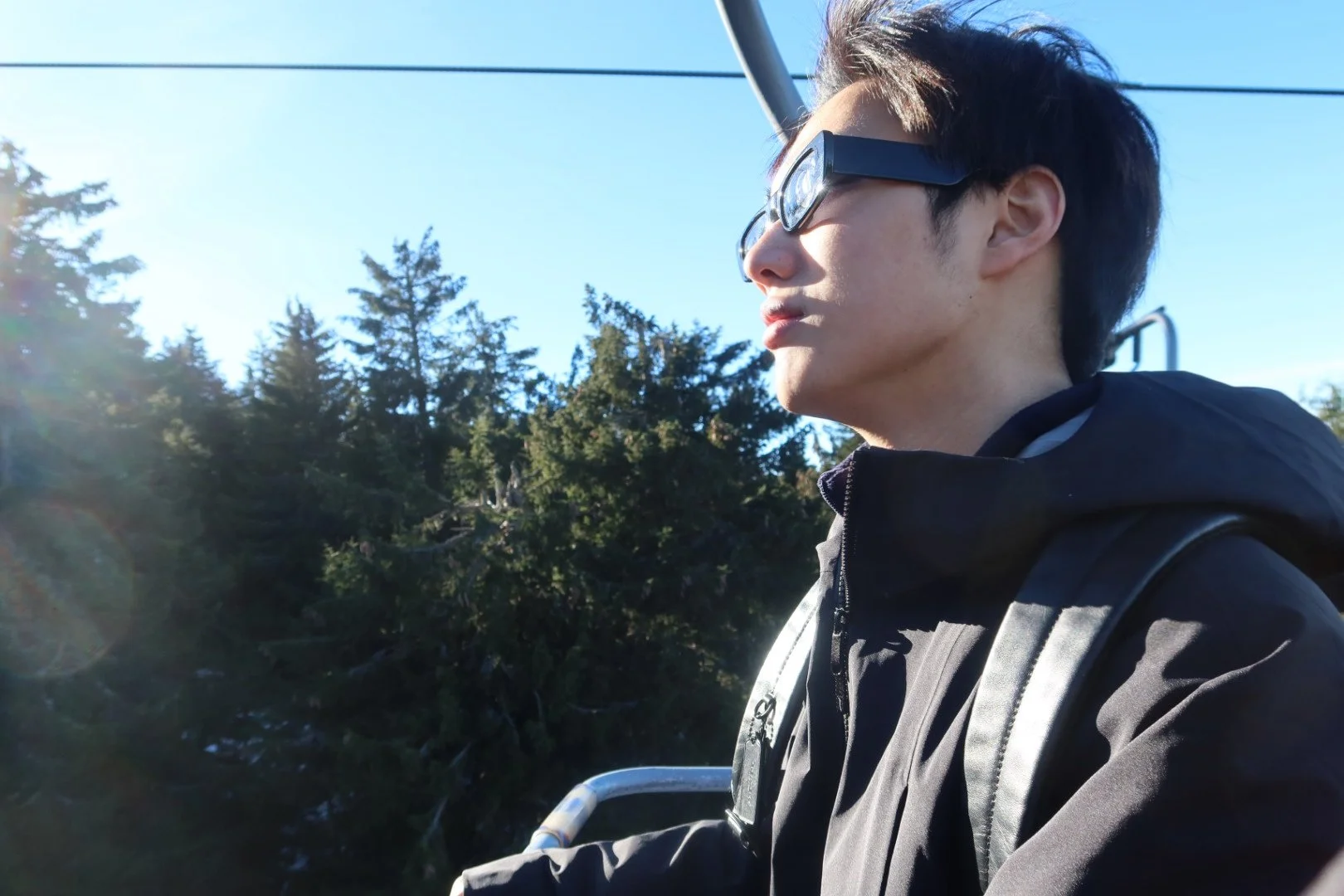 Young man wearing sunglasses and a black jacket outdoors with trees and blue sky in the background.