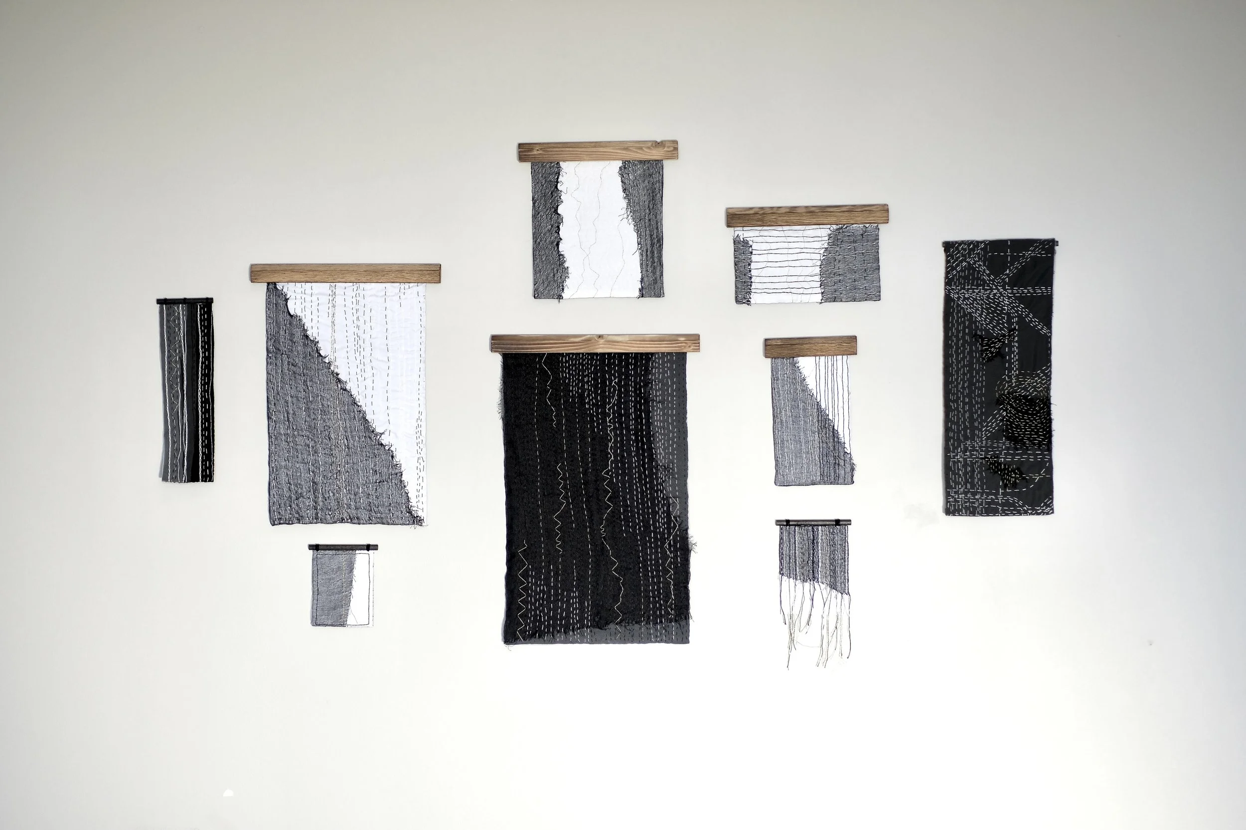Display of seven textile art pieces hanging on a white wall, each with black, white, and gray patterns, some with layered textures and wooden supports.