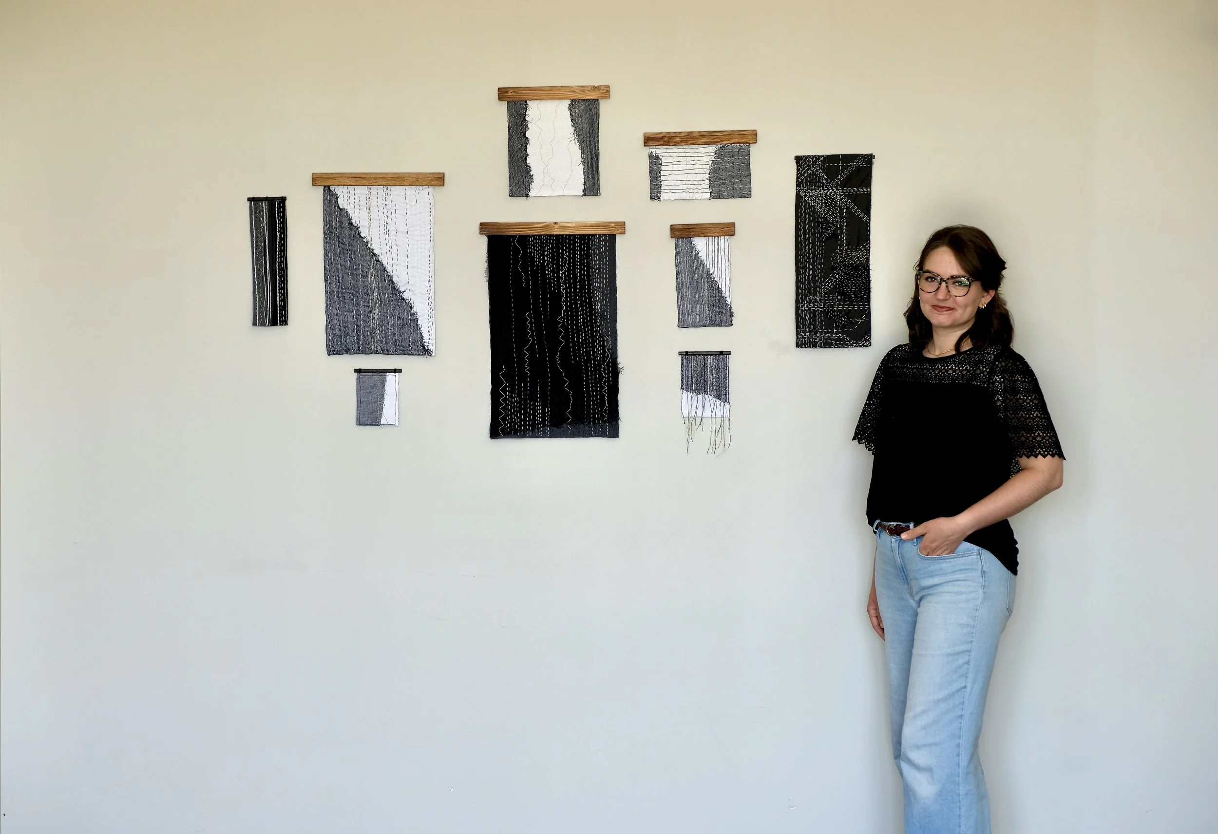 Artist Sarah Kirby, standing beside abstract black and white textile art pieces on a pale yellow wall.