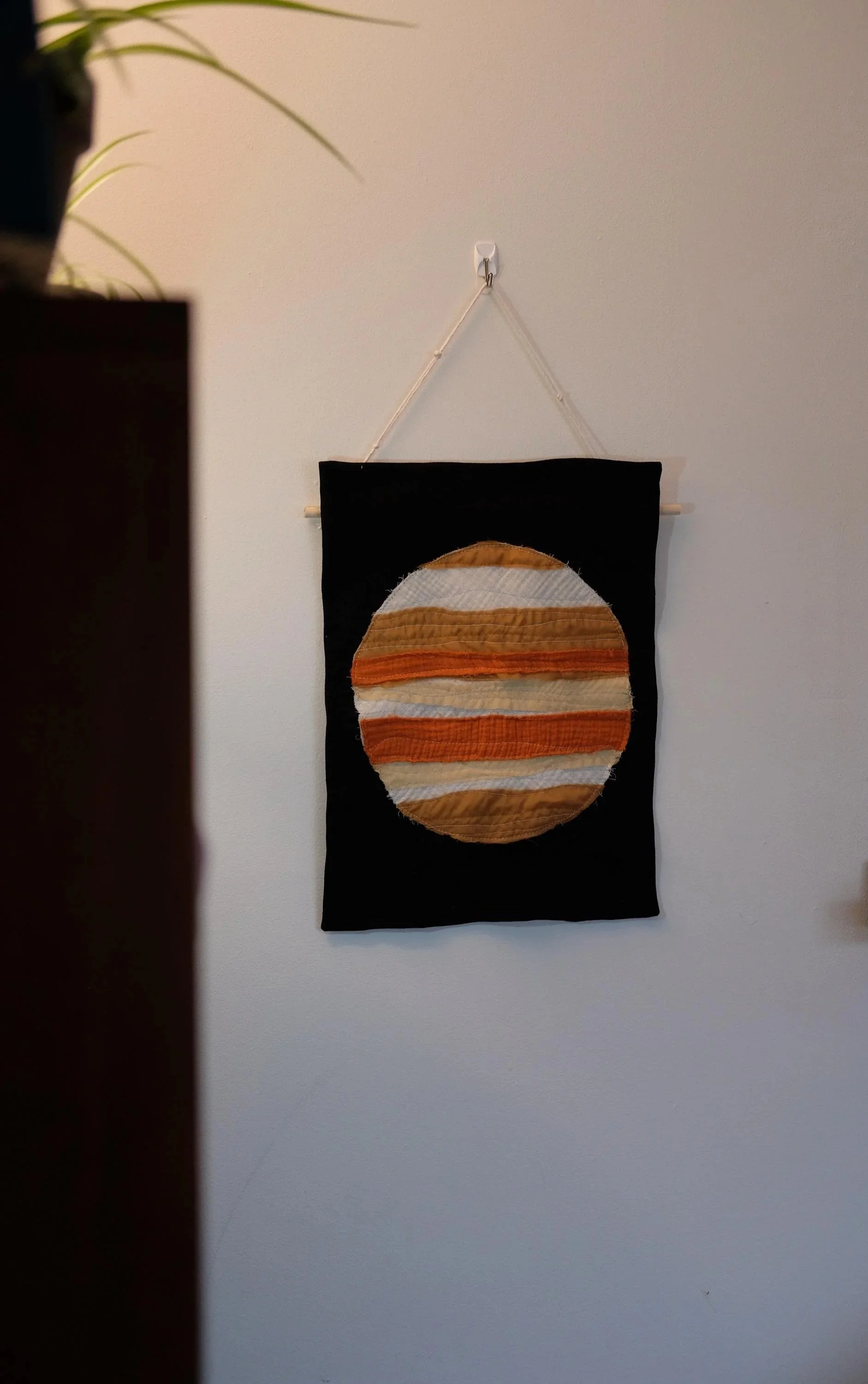 A decorative wall hanging featuring a circular, striped pattern in shades of orange, cream, and brown, mounted on a black fabric background and hanging from a white hook on a white wall.