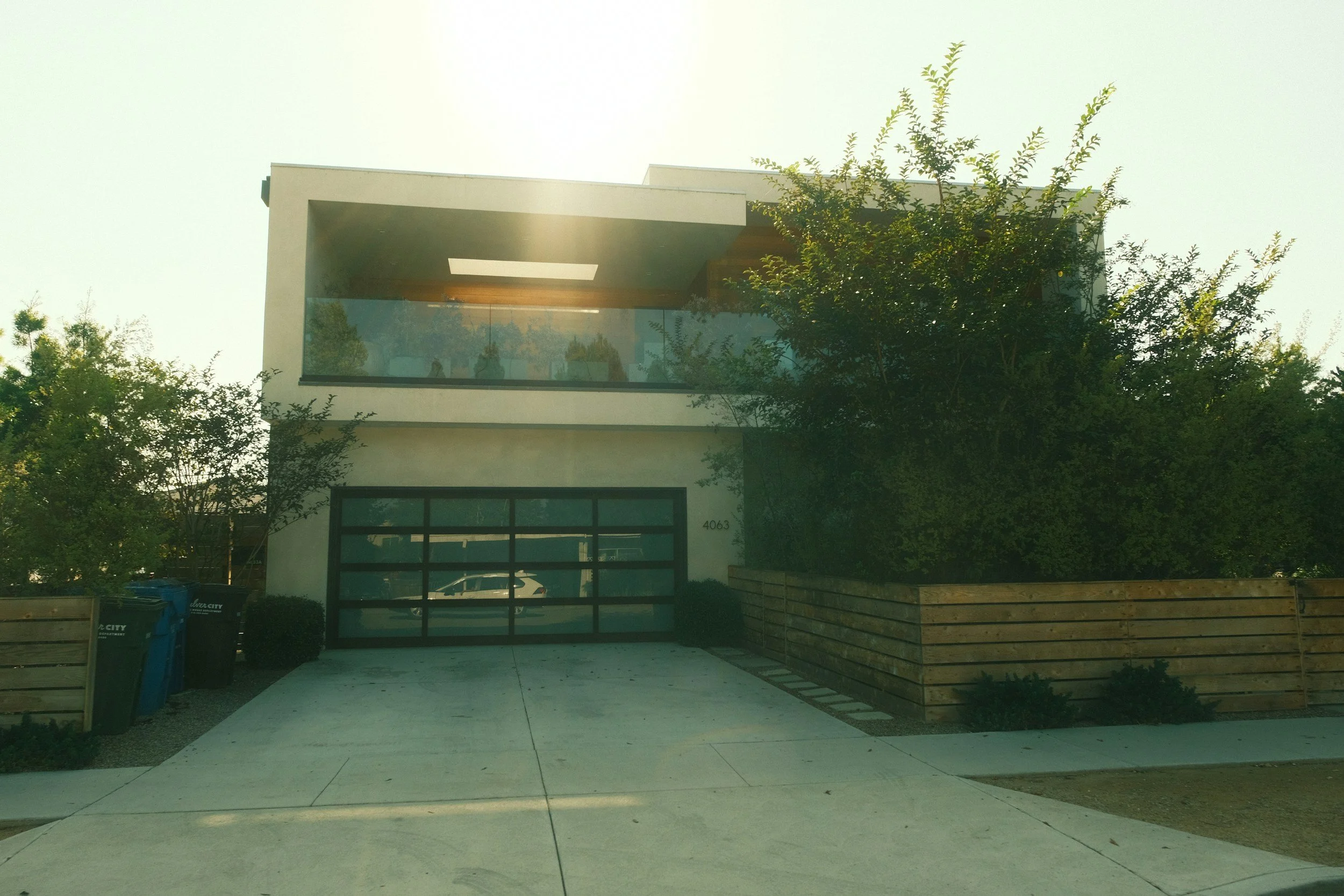 Modern white two-story house with a glass balcony, surrounded by trees, driveway, and wooden fence.
