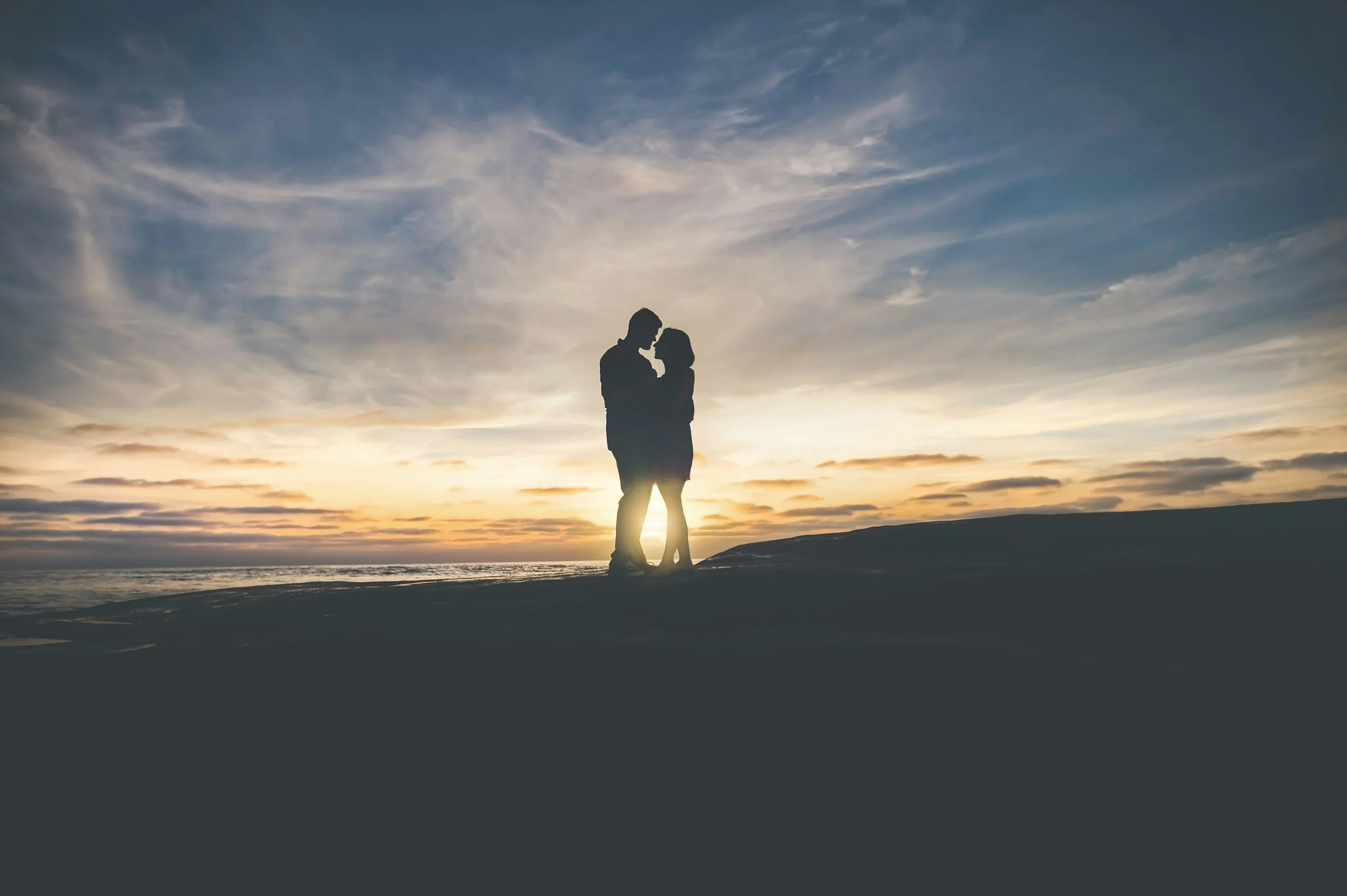 Silhouette of a couple embracing at sunset on a beach with a cloudy sky overhead.