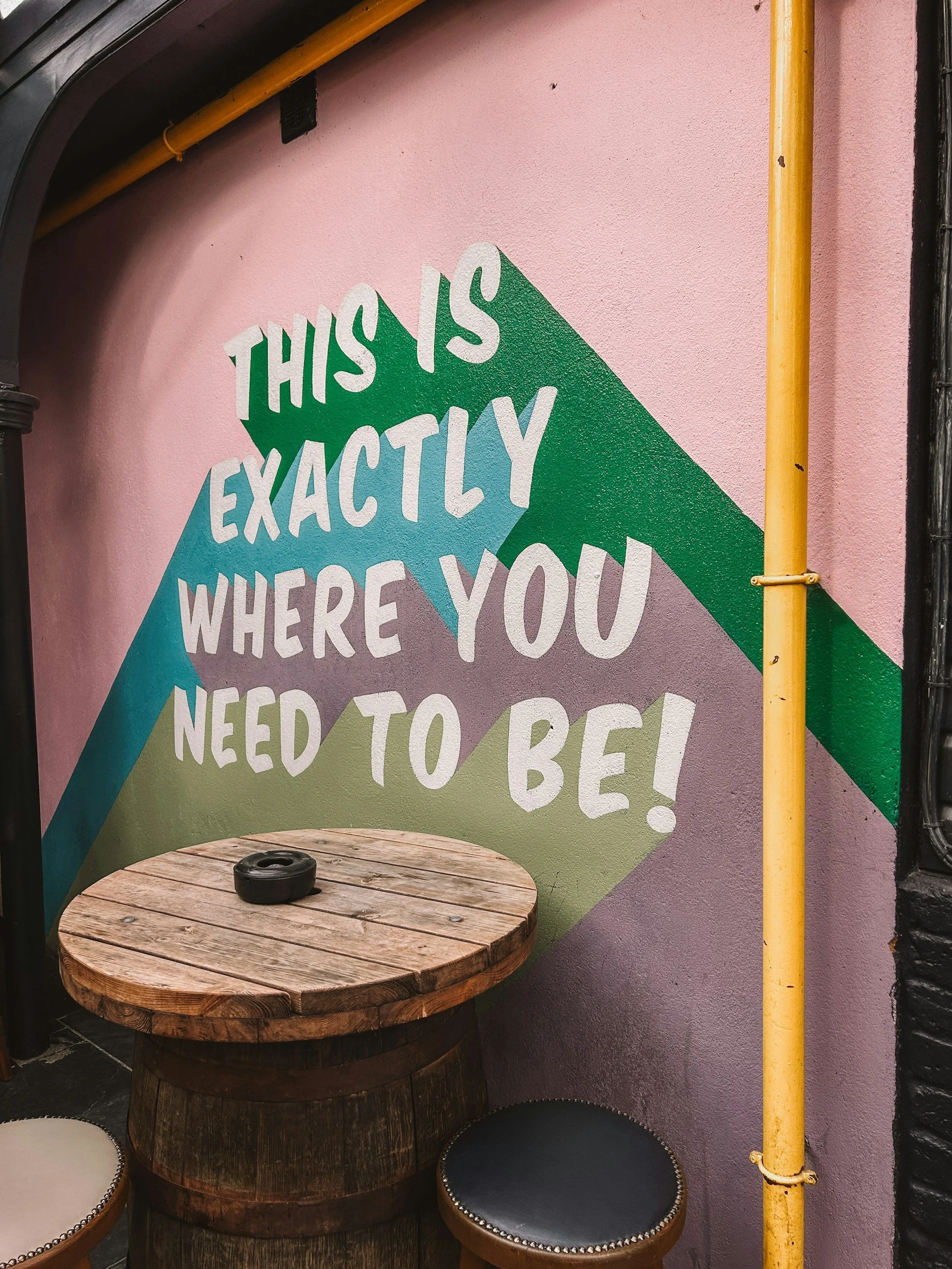 A mural on a pink wall with the message "This is exactly where you need to be!" painted in white and colorful letters. A round wooden table with a black ashtray and two stools with cushion seats are in front of the mural. A yellow pipe runs vertically along the right side of the image.
