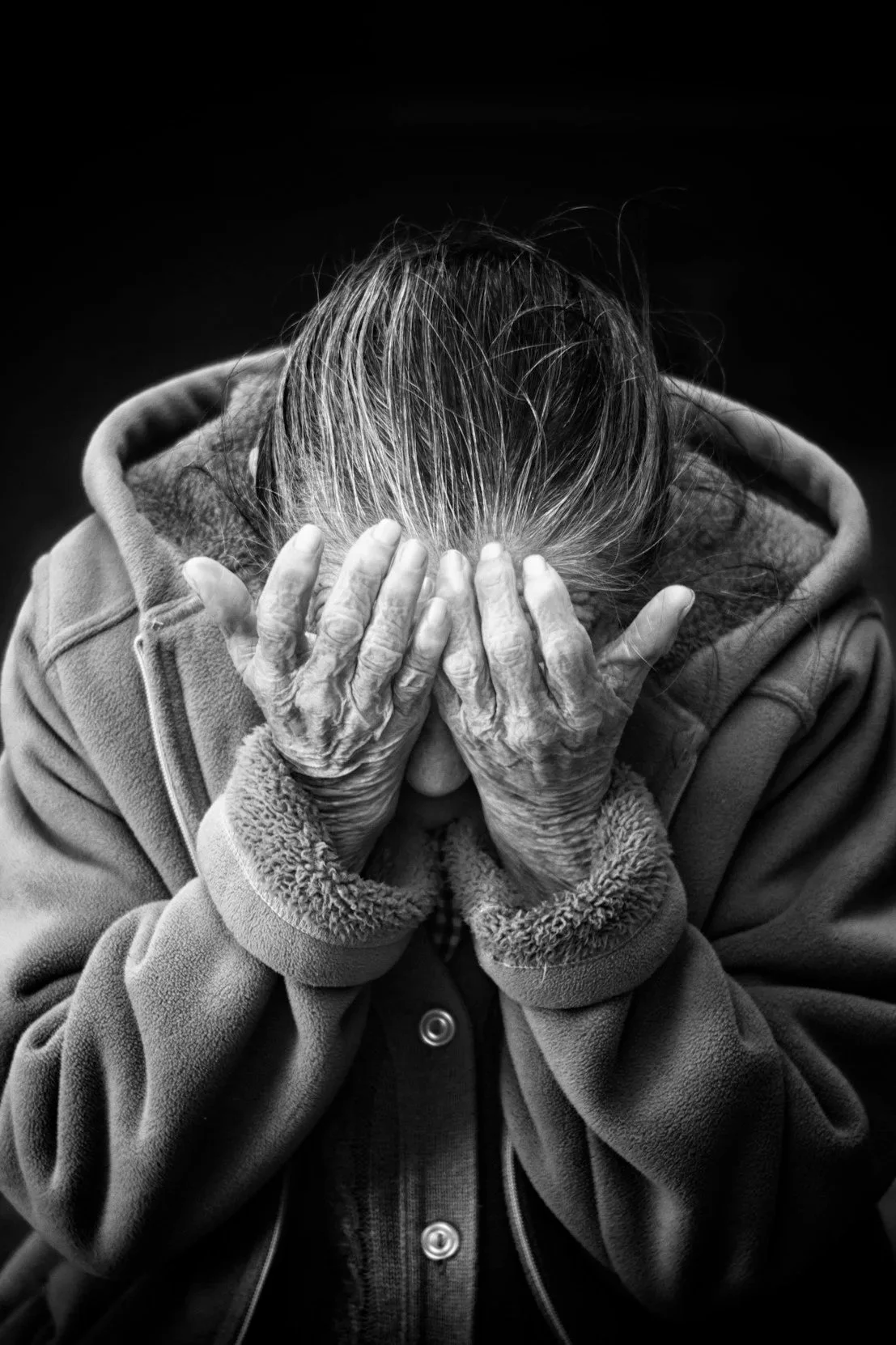 An elderly woman with gray hair covers her face with her hands, wearing a coat with fur-lined cuffs, against a black background.