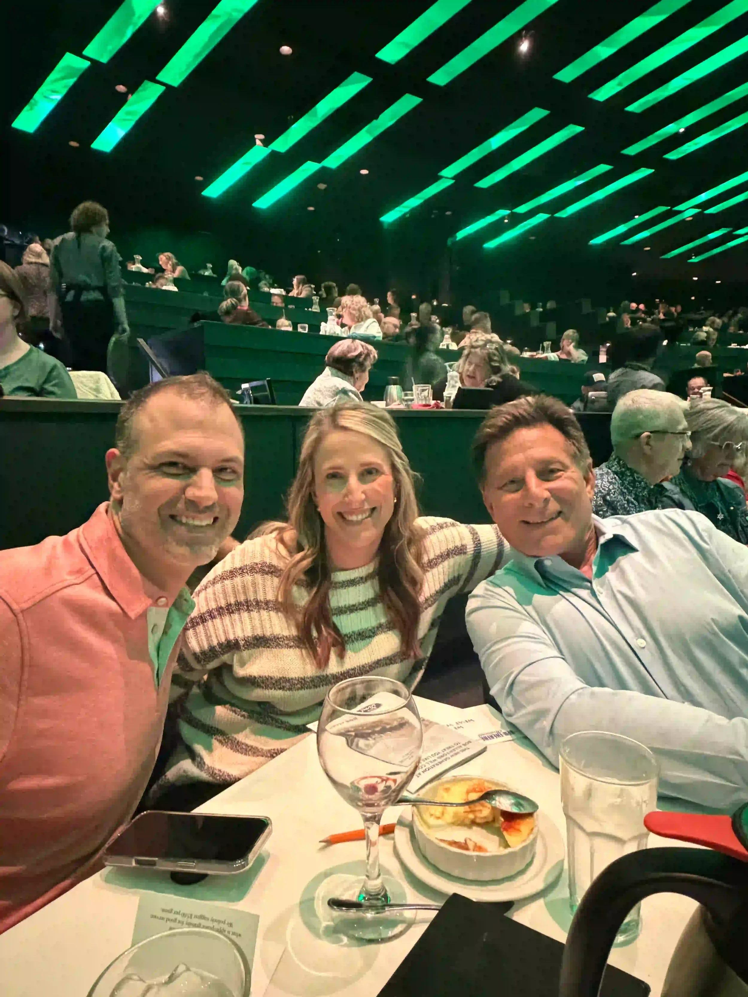 Inside a crowded restaurant with green ceiling lights, three smiling people sitting at a table with wine glasses, a plate of pizza, and a smartphone in front of them.