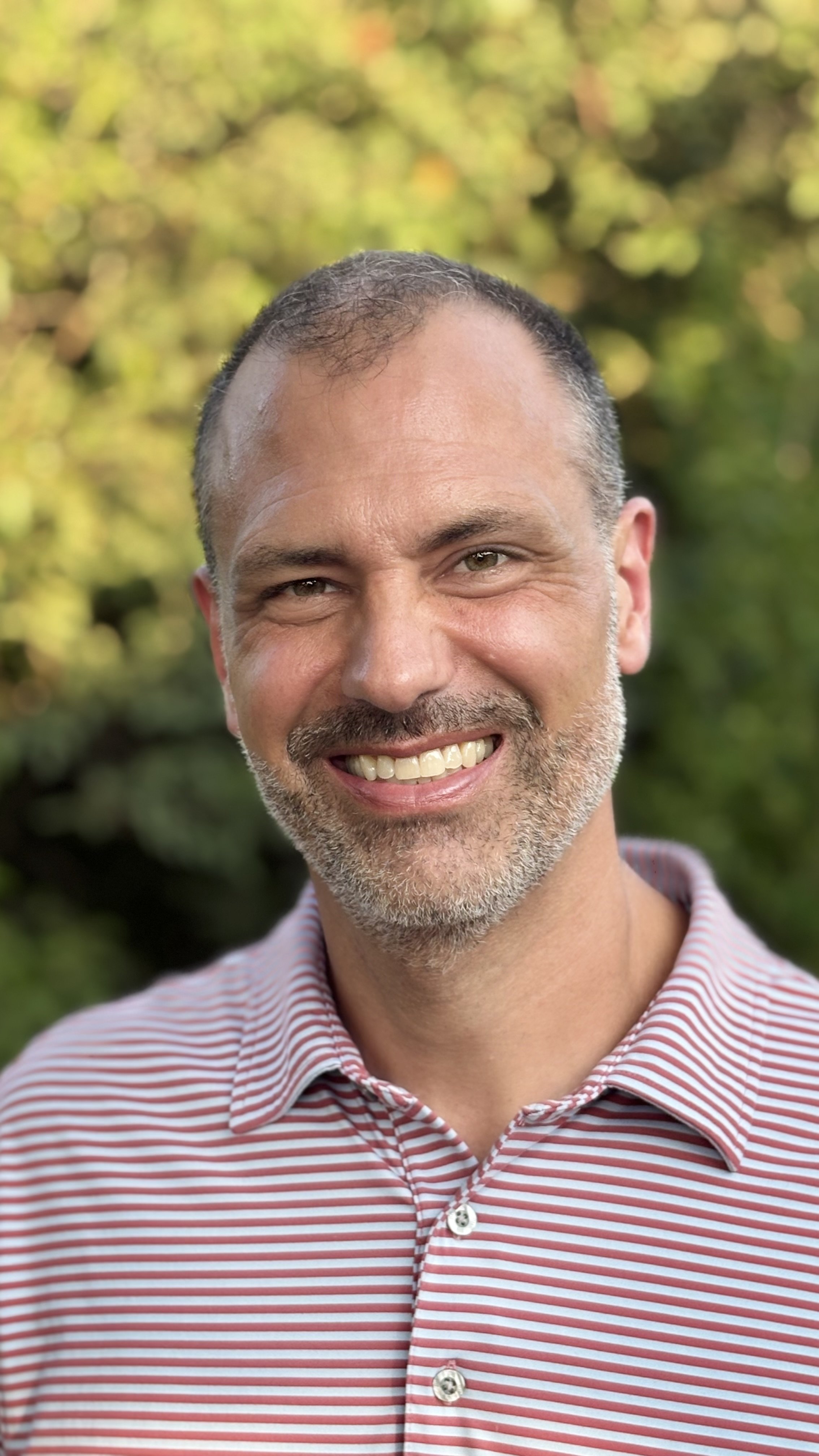 Close-up of a smiling man wearing a red and white striped collared shirt, with a blurred green foliage background.