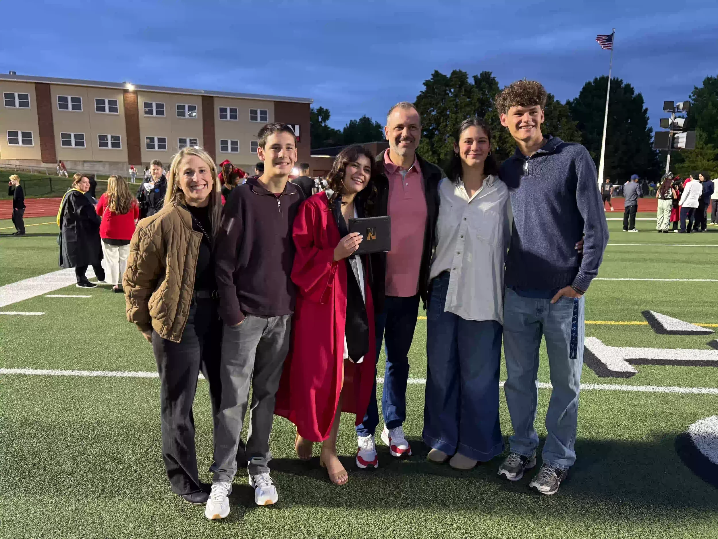 Group of six people standing on a sports field at dusk, smiling for a photo, with a building, trees, and an American flag in the background.