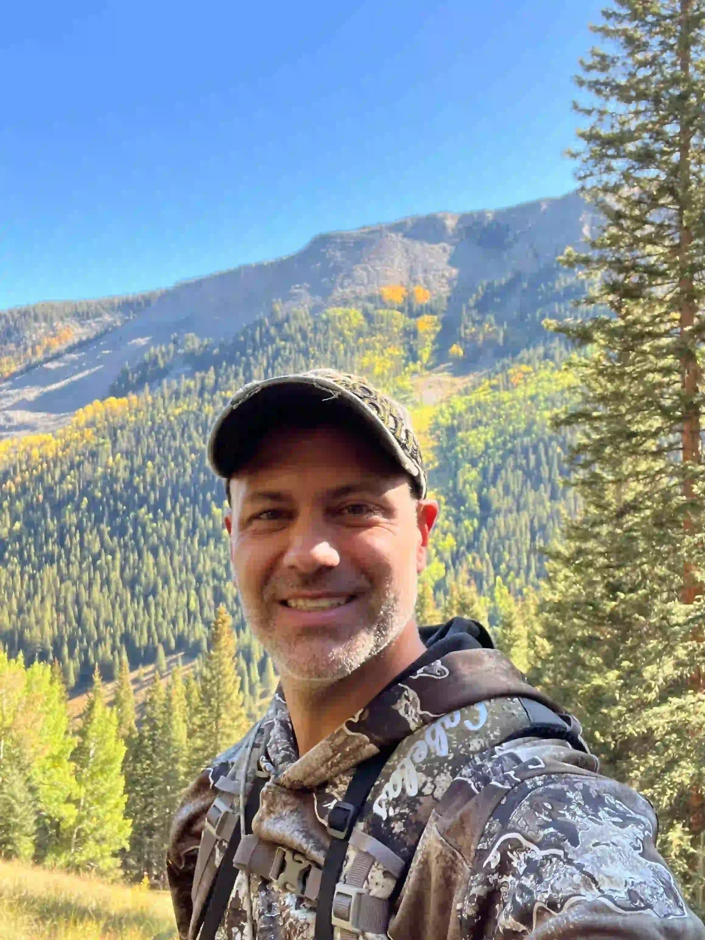 A man smiling outdoors in a forested mountain area with vibrant fall foliage, wearing a camouflage jacket and a cap.