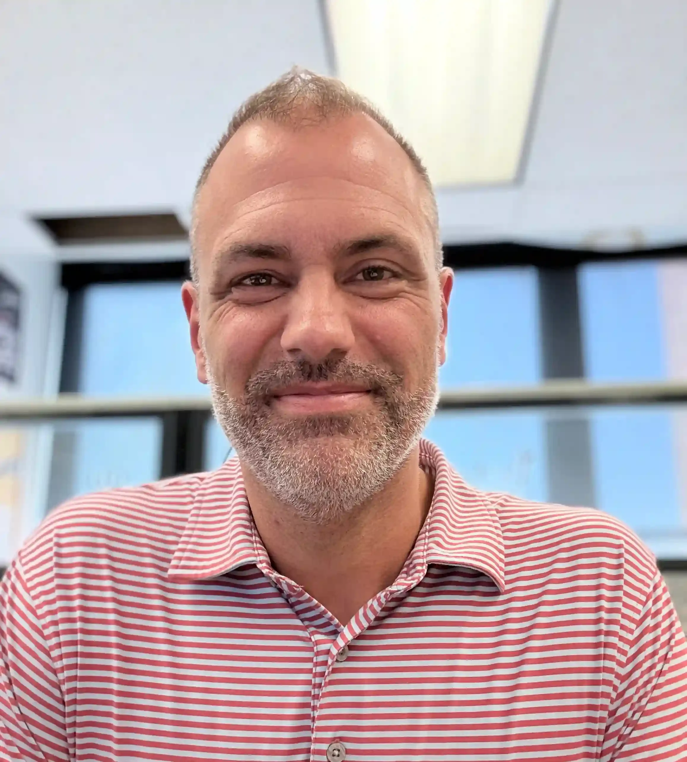 Close-up of a middle-aged man smiling, wearing a red and white striped collared shirt, with a background of windows and a light-colored ceiling.