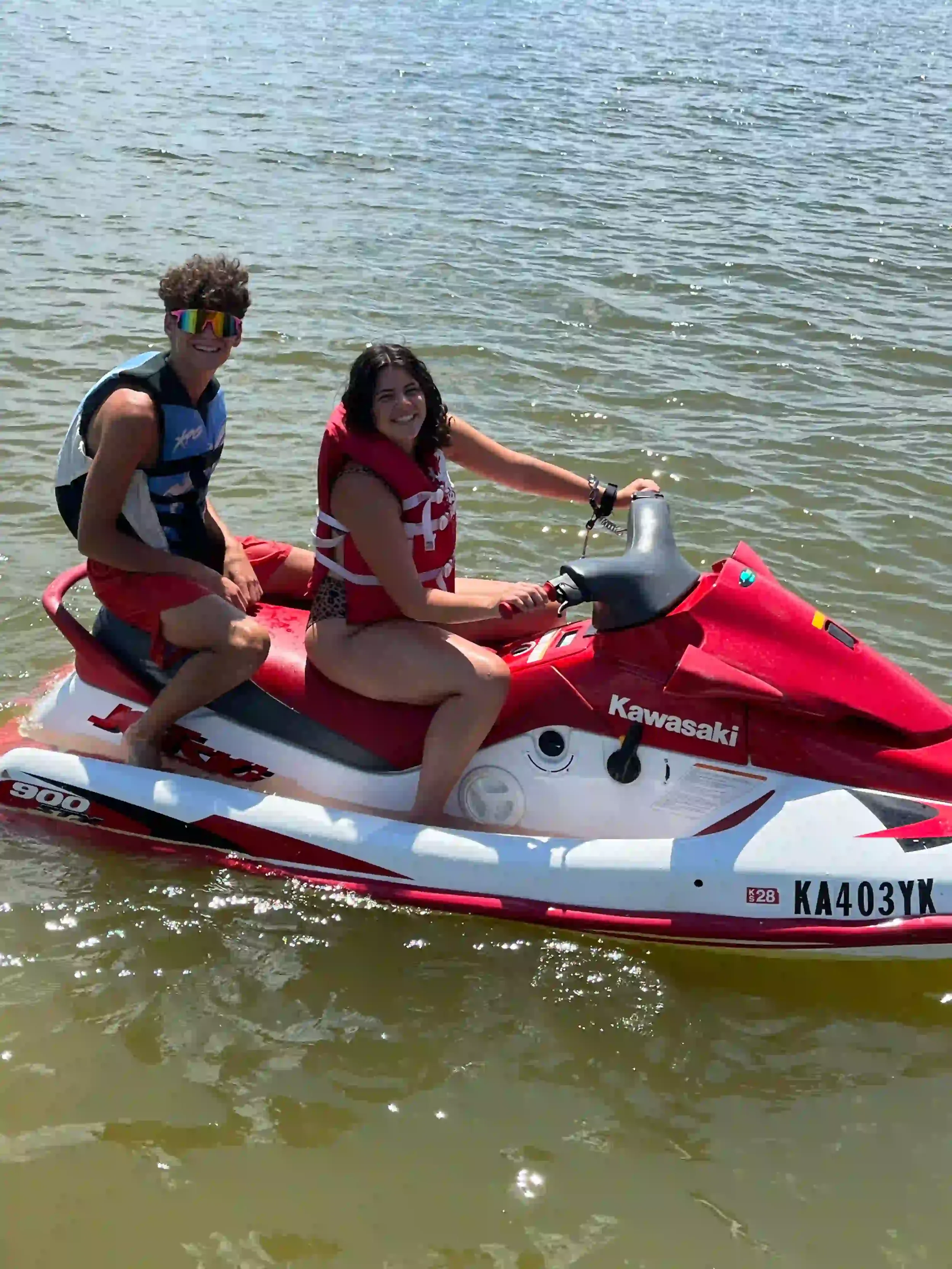 Two young people riding a red and white Kawasaki jet ski on a body of water, both wearing life jackets and smiling.