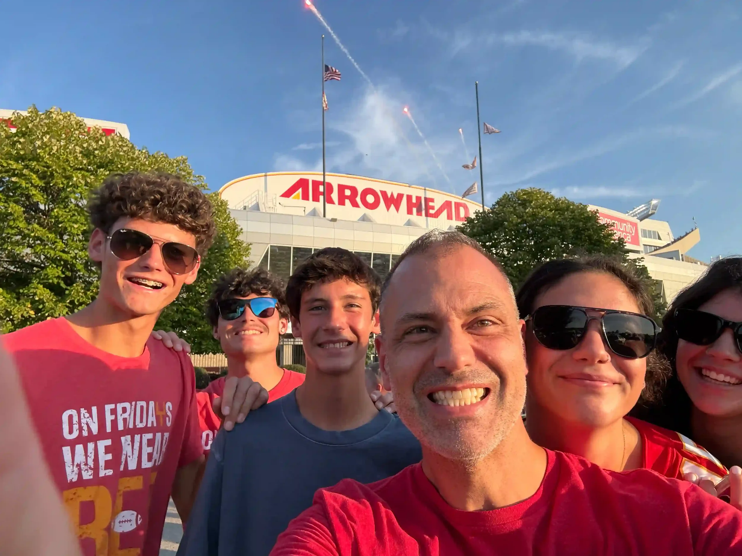 Group of people taking a selfie in front of Arrowhead Stadium with fireworks and American flags in the sky.