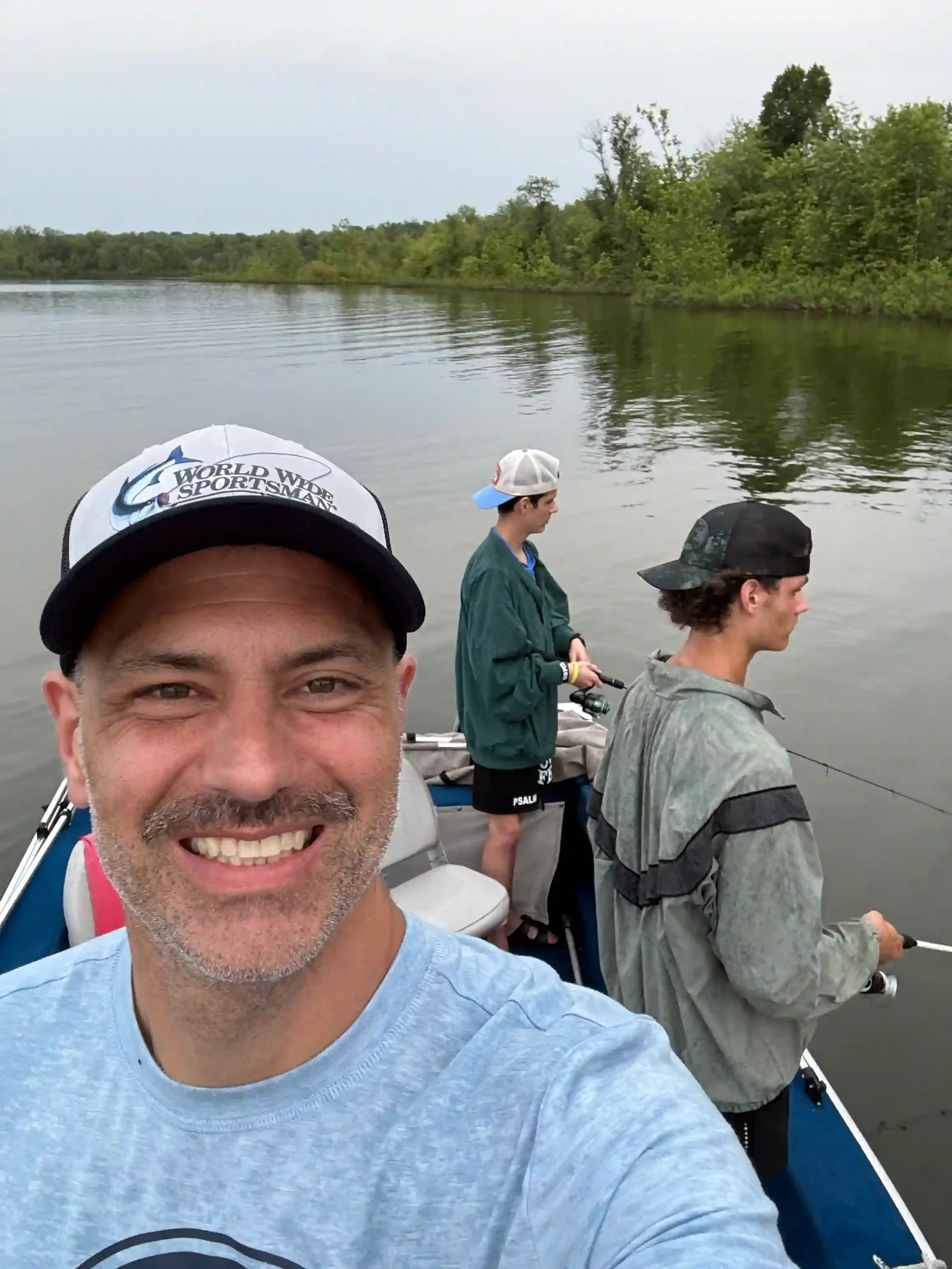 Smiling man taking selfie on boat with three young men fishing in the background, water and green trees visible.
