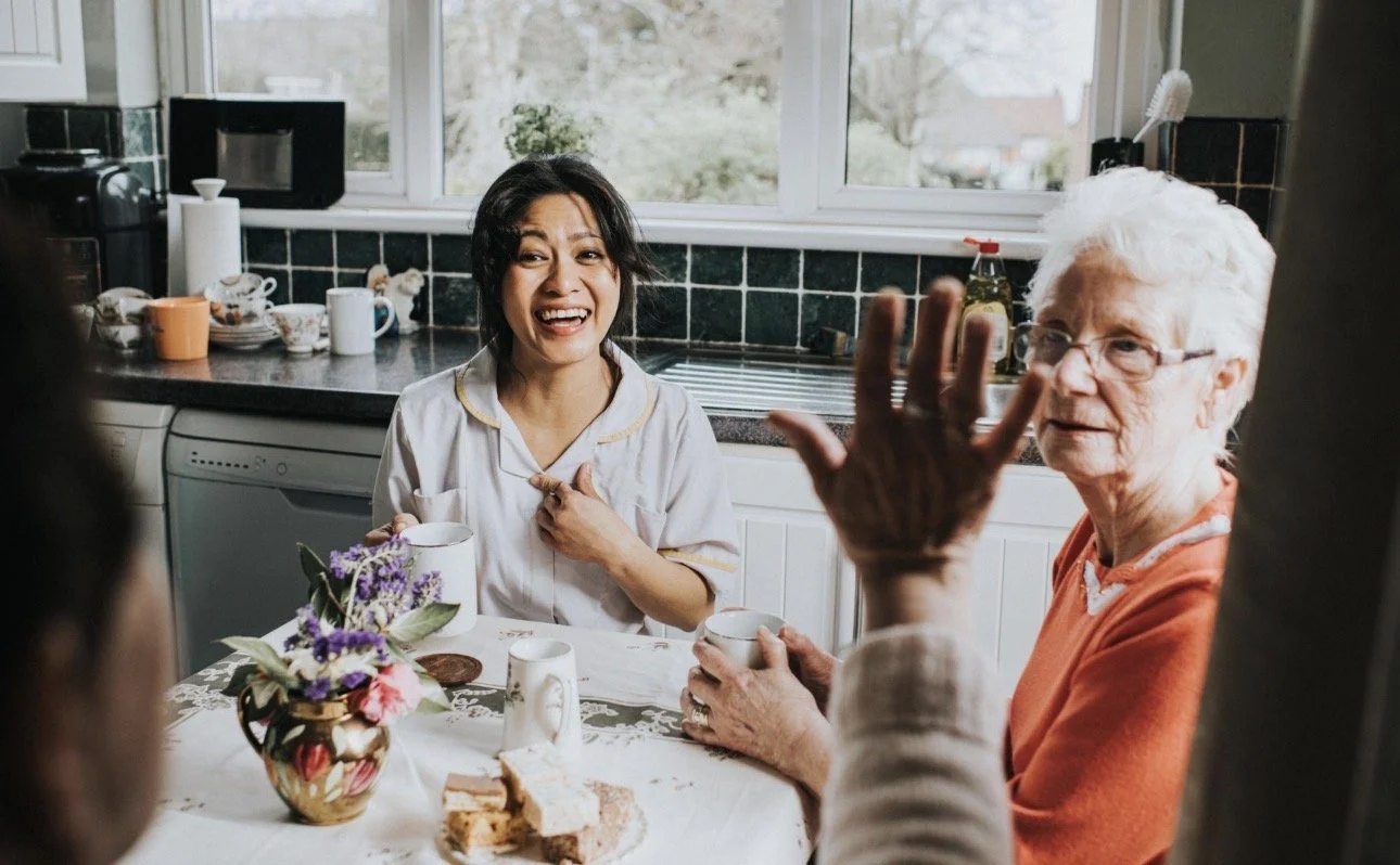 Four women are sitting around a kitchen table, engaging in conversation. One woman with short white hair and glasses is waving her hand and talking, while another woman with black hair is smiling and holding a mug. The table has a vase with flowers, teacups, a plate with cake slices, and other items. The kitchen has a window showing an outdoor scene.