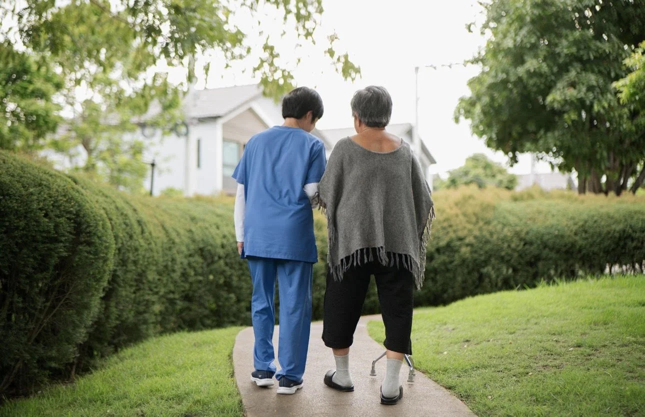 A young woman and an elderly woman are walking on a sidewalk in a residential area, surrounded by greenery and trees