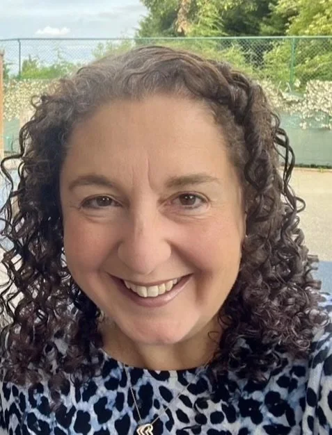 Close-up photo of a woman with curly hair smiling outdoors in front of a tennis court and green trees.
