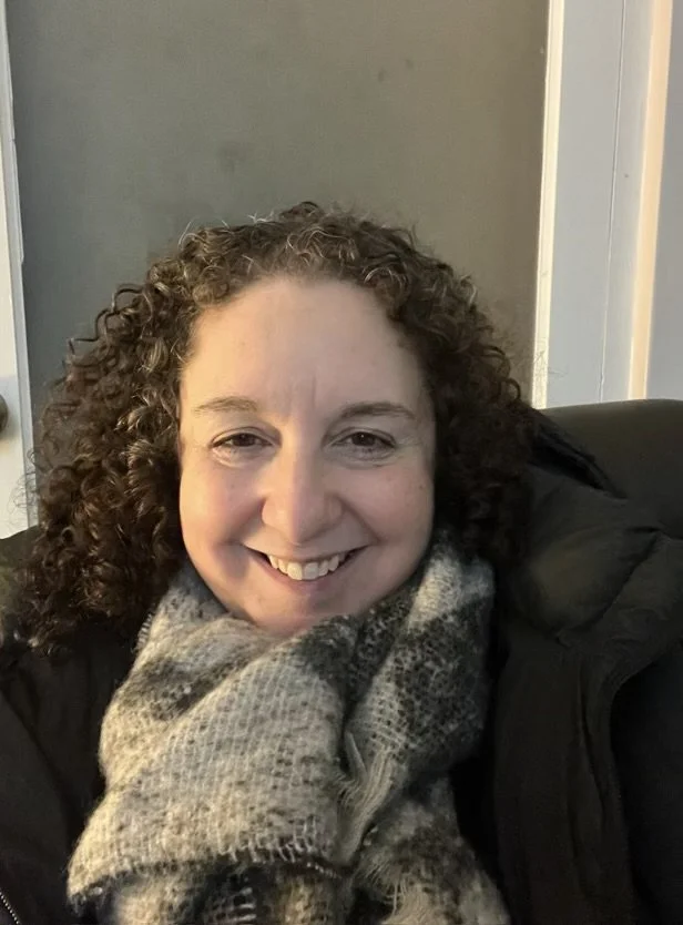 A woman with curly hair smiling at the camera, wearing a scarf and a black jacket, sitting in a cozy indoor setting.