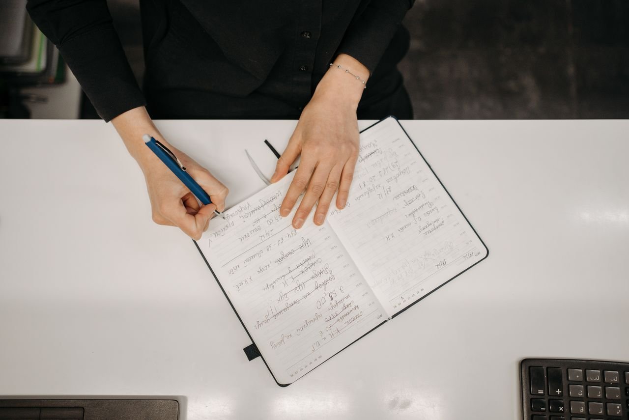 A person writing in a notebook on a white desk, with a pen in their right hand and the notebook in front of them.