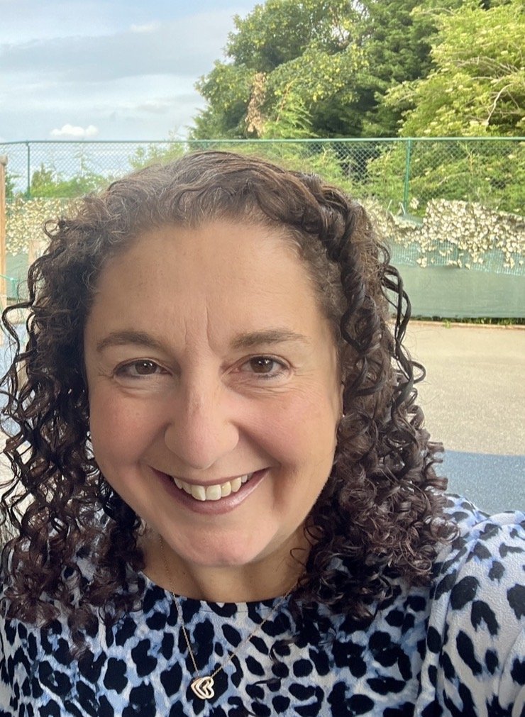 Close-up of Sarah Greenfield, founder of Green Thread smiling outdoors, wearing a black and white animal print top and a heart-shaped necklace, with a tennis court and country background.