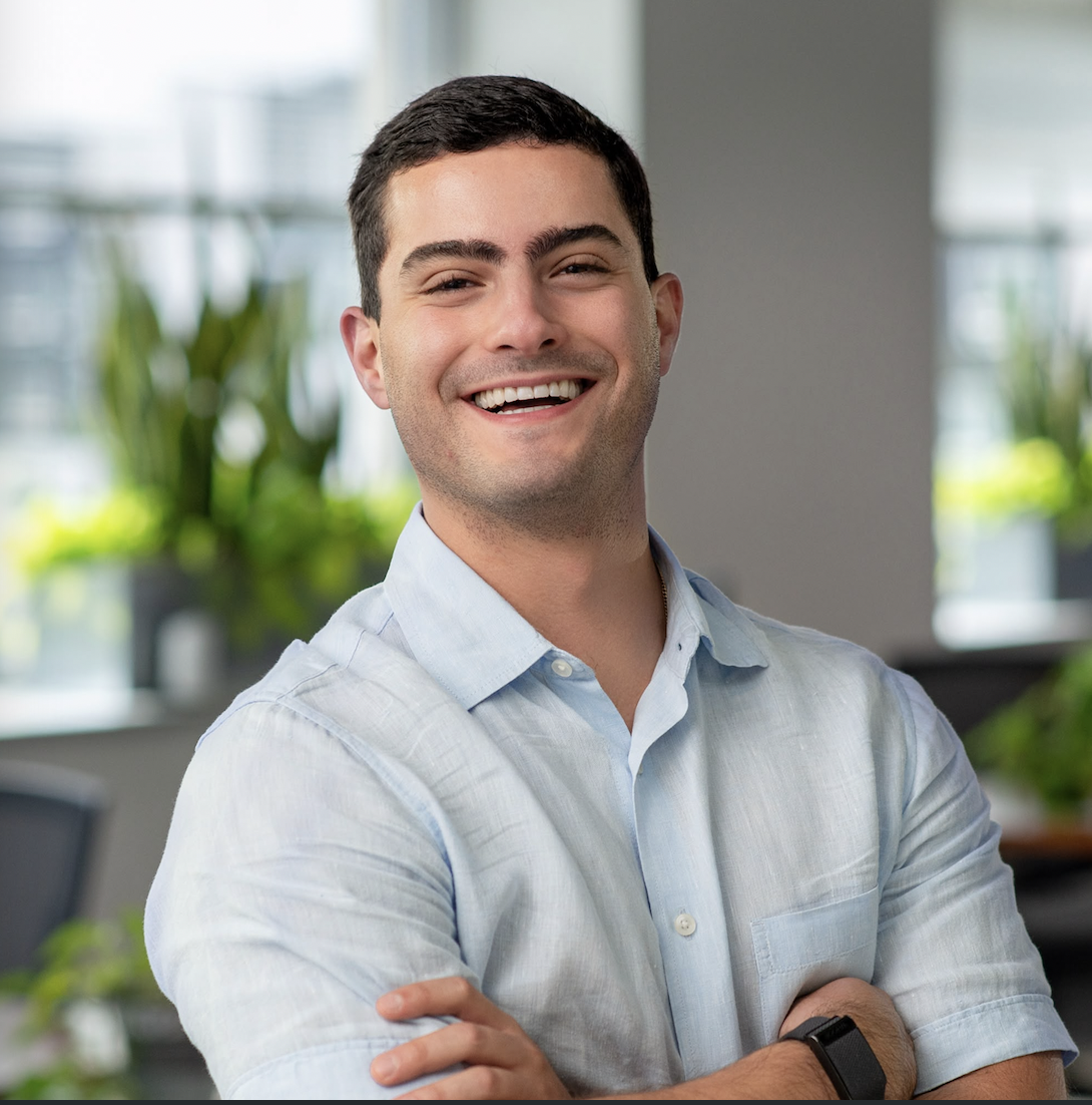 A young man with short dark hair smiling, wearing a light blue button-up shirt, in an office setting with plants in the background.
