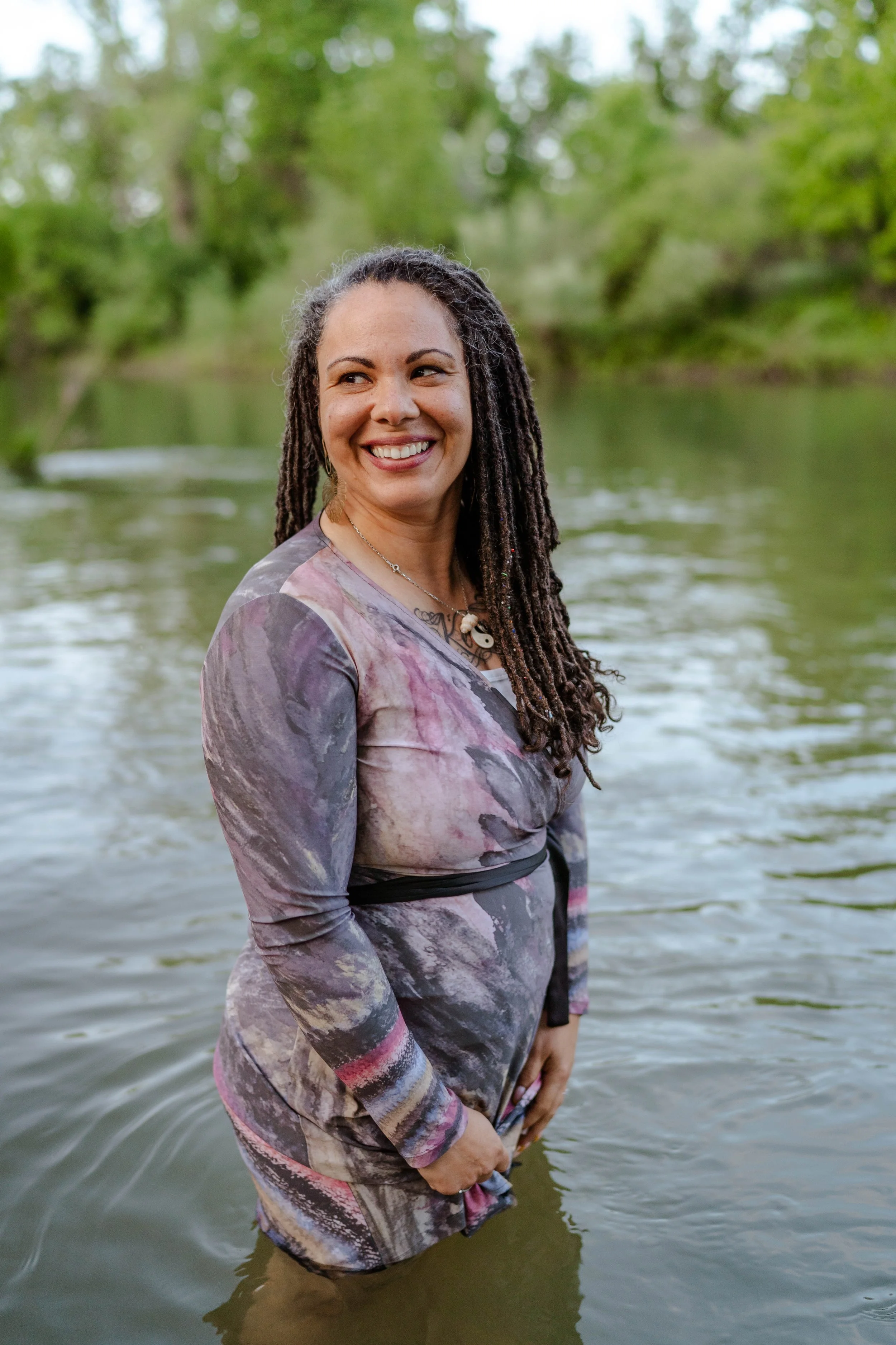 A woman with long dreadlocks smiling while standing waist-deep in a river, surrounded by green trees in the background.