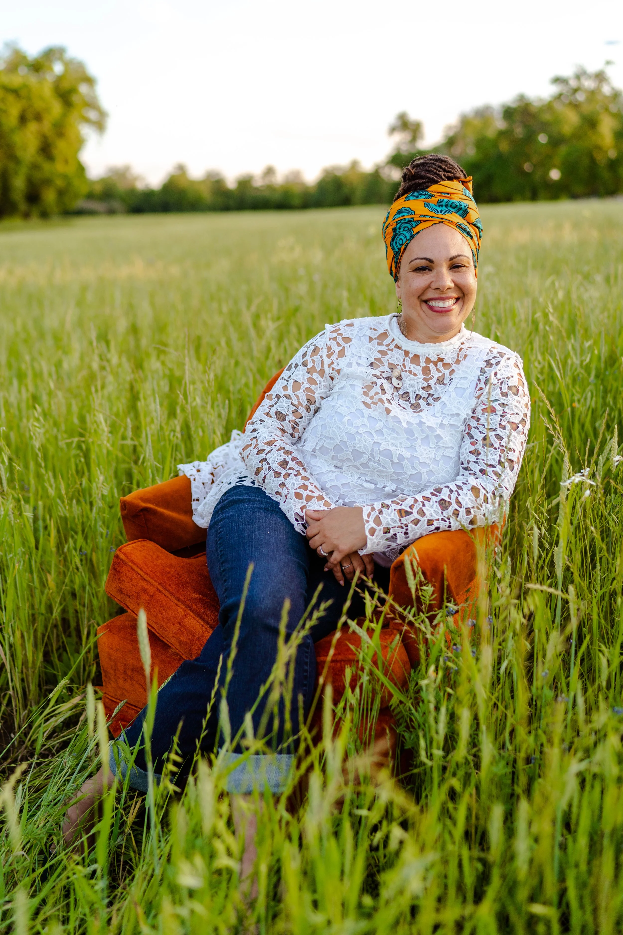 A woman with a yellow and blue patterned headwrap smiling while sitting on an orange velvet armchair outdoors in a grassy field with trees in the background.