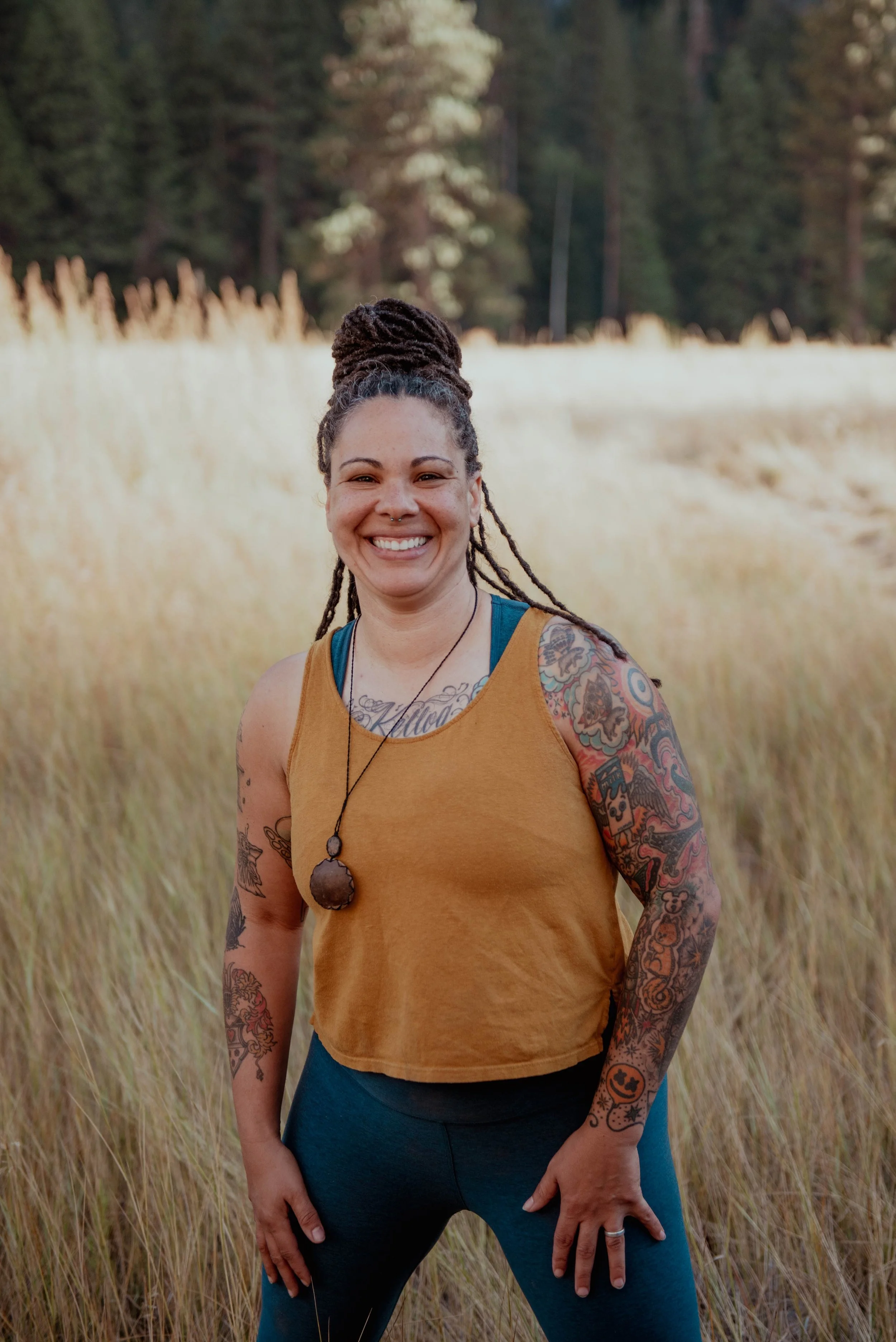 A smiling woman with dreadlocks tied up in a bun, wearing a mustard yellow sleeveless top and dark leggings, standing in a field of tall grass with trees in the background.