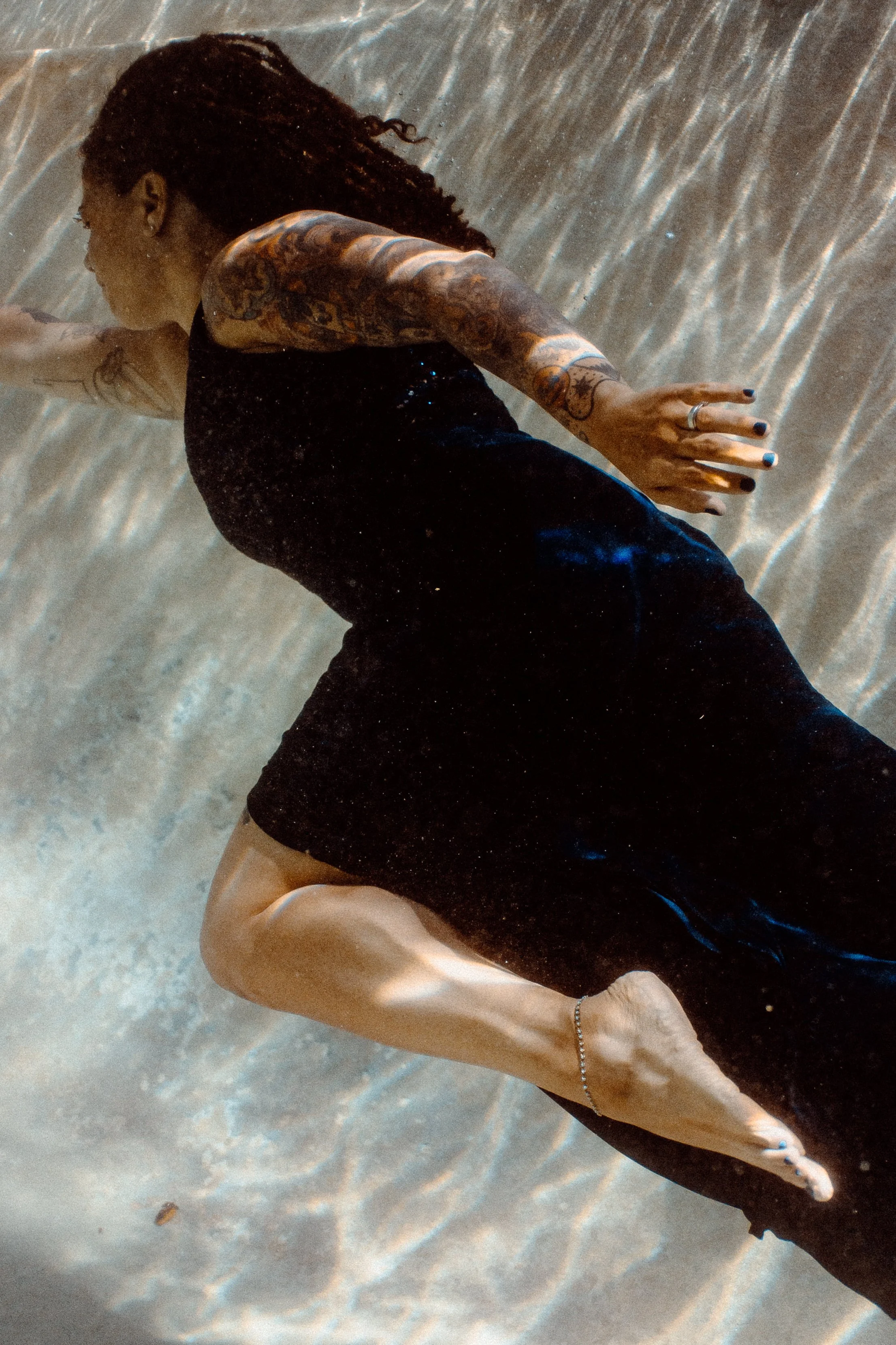 A person lying on a sand surface, with visible tattoos on their arm and hand, wearing a black shirt and black painted nails, with an arm extended and a finger pointing outward.