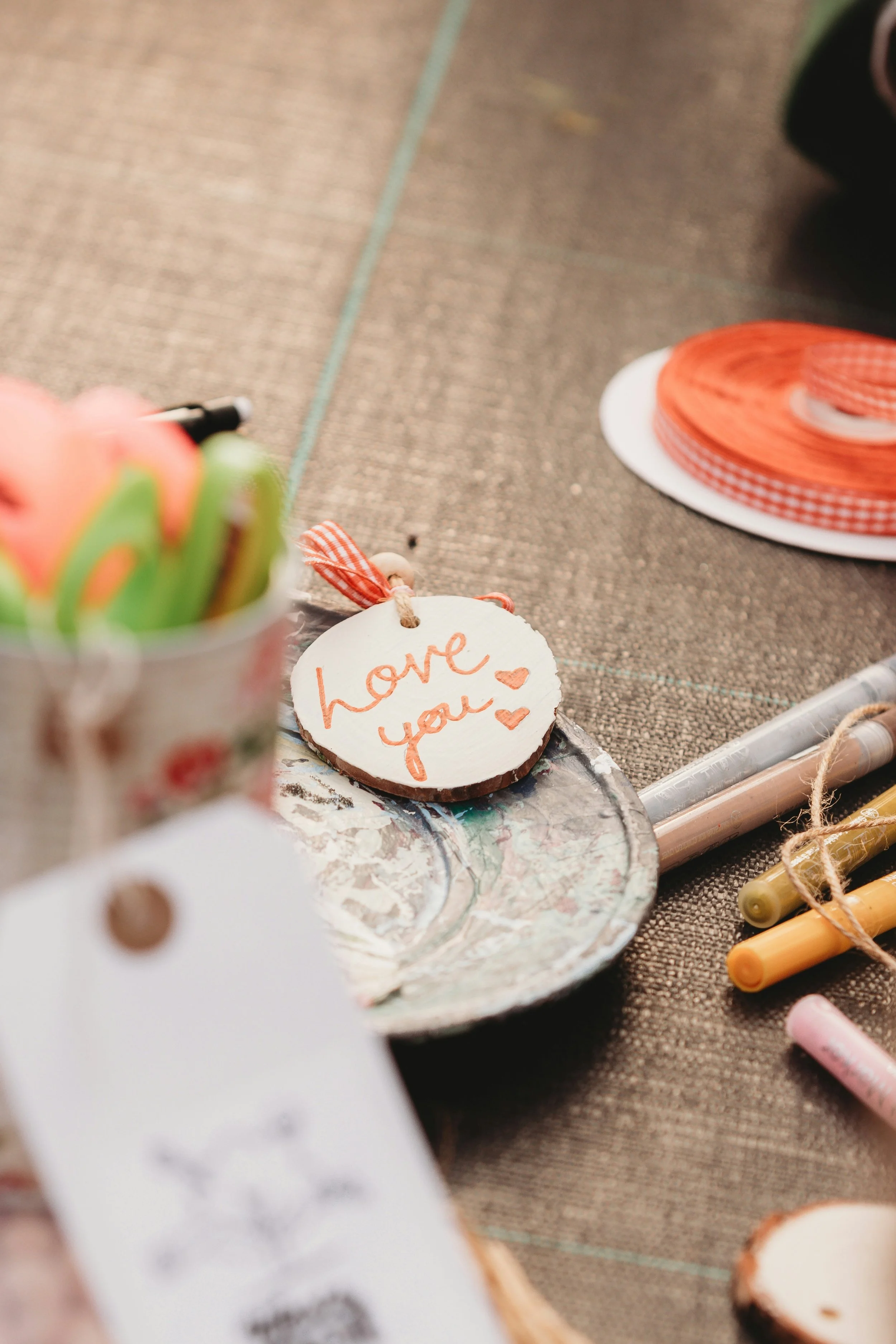 Close-up of a decorative wooden slice with handwritten orange text that says "love you" and small heart drawings, surrounded by crafting materials including ribbons, yarn, and markers on a textured surface.