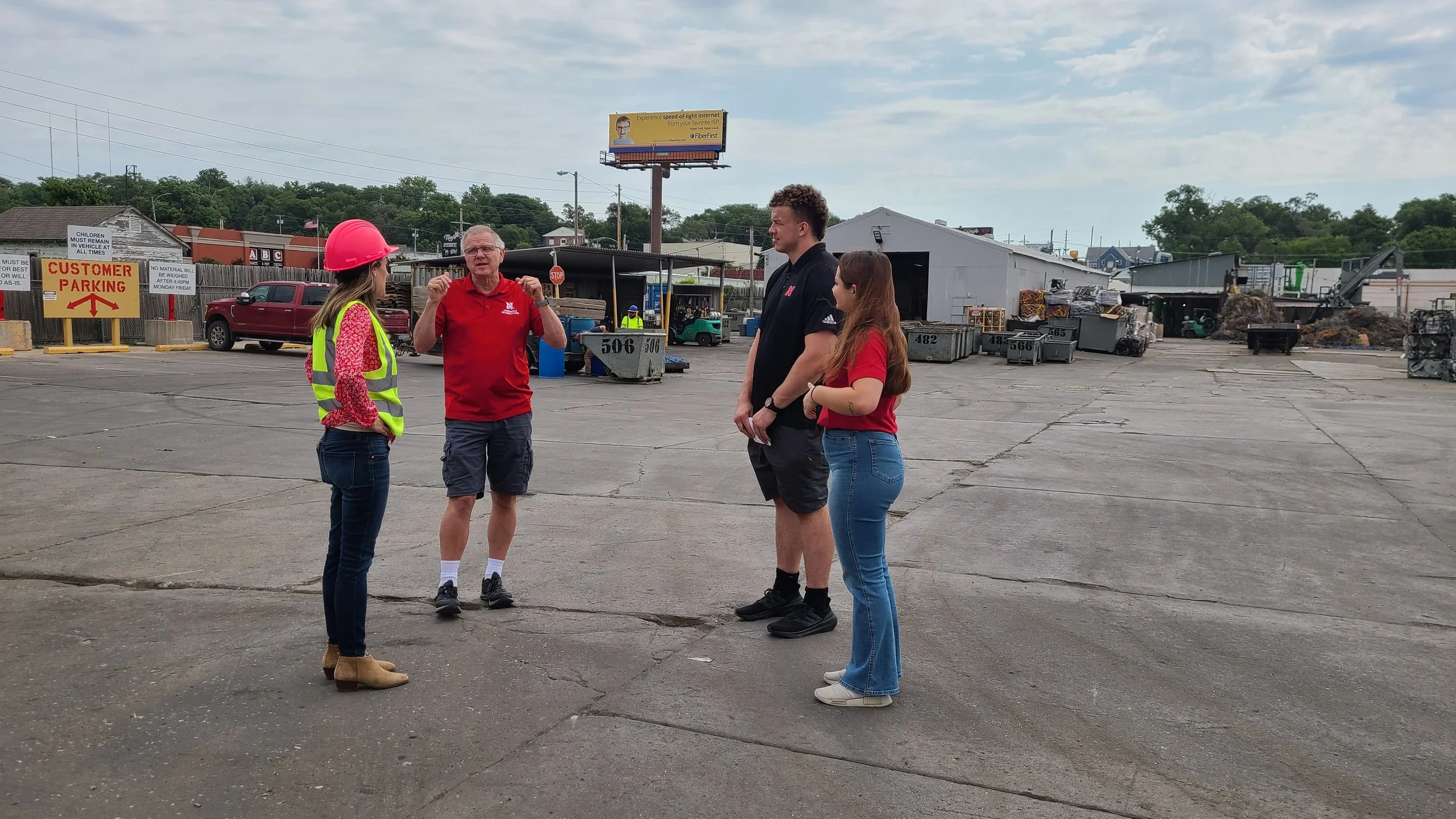 Four individuals engaged in conversation outside in a parking lot, with industrial and residential buildings in the background. One woman in a pink safety helmet and yellow reflective vest is speaking to an older man in a red shirt, while two younger individuals, a man and a woman, listen. All are casually dressed, and the scene appears to be a discussion or orientation.