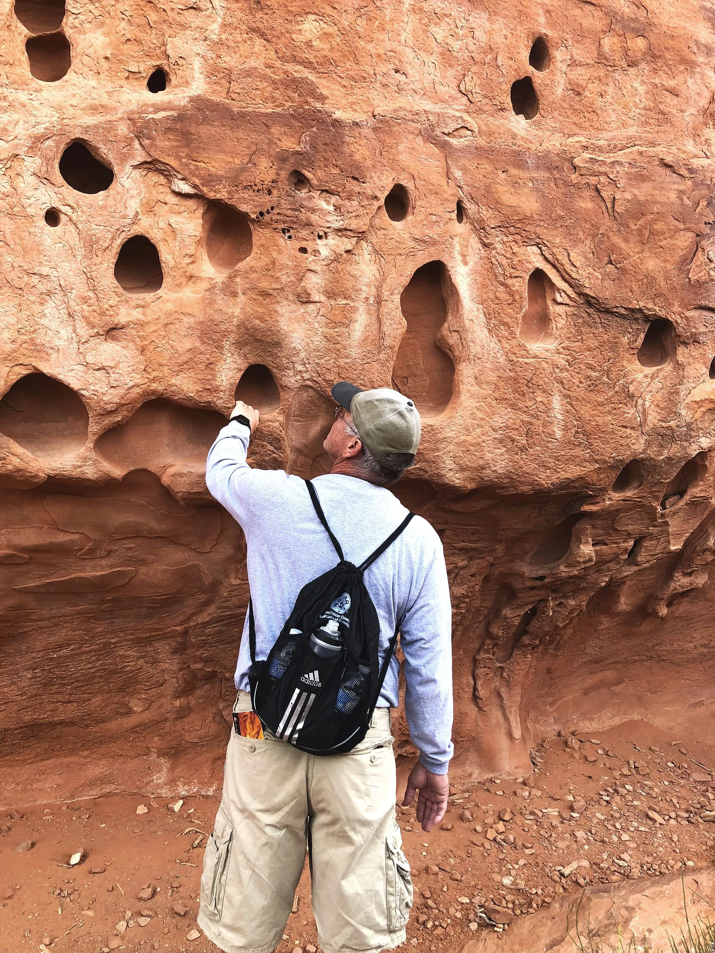 A man pointing at a large red rock formation with numerous small holes and cavities. He is wearing a light gray hoodie, khaki cargo shorts, a gray cap, and has a black backpack with a water bottle attached.