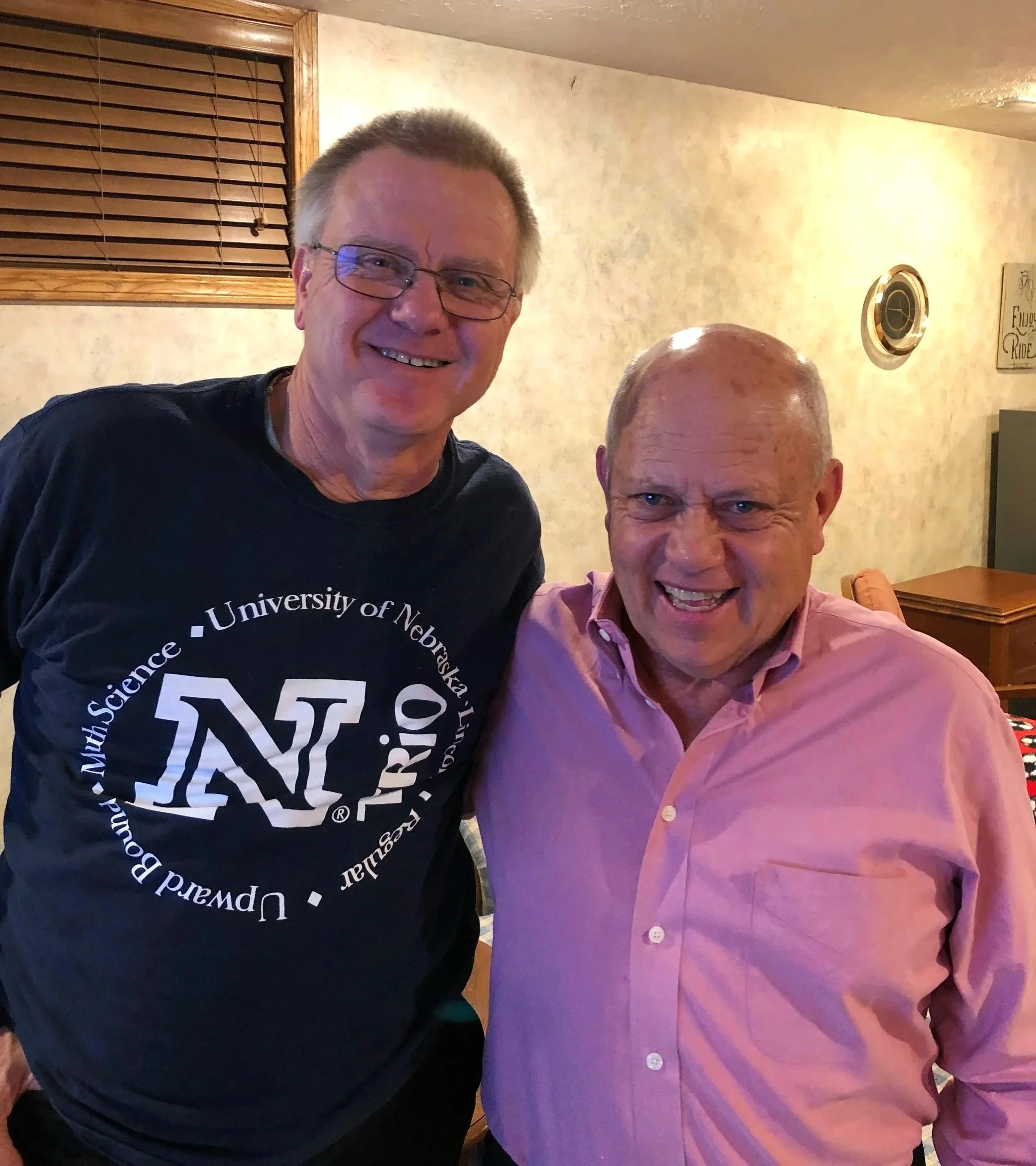 Two smiling men stand close to each other, with parts of their faces and shoulders visible, in an indoor setting with beige walls, wooden window blinds, and a decorative sign in the background. One man wears a black shirt with white text related to t