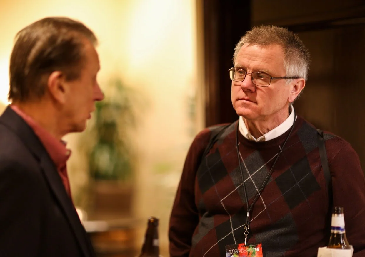 Two men having a serious conversation at a social gathering, one facing away from the camera, the other wearing glasses and a sweater with a badge, drinks in hand.