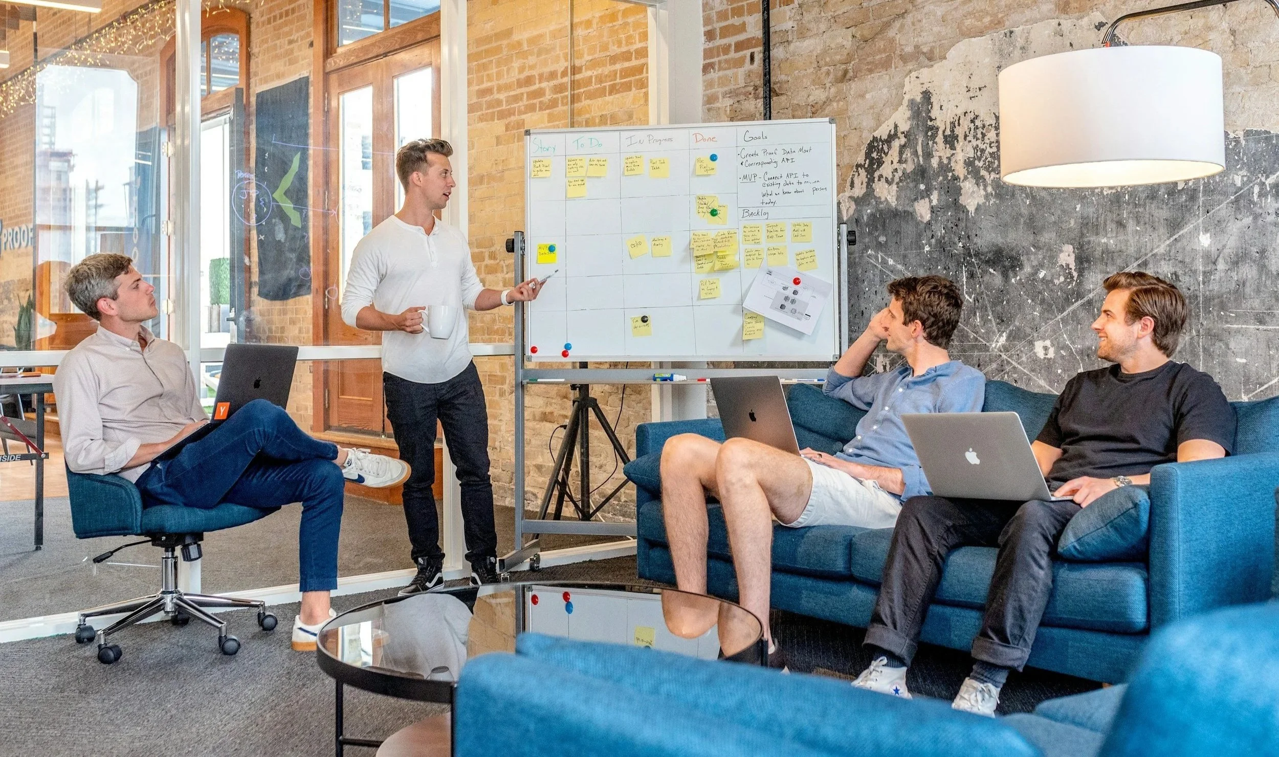 Group of five young men in a modern office or meeting space. One man stands near a whiteboard with sticky notes and notes written on it, gesturing as he speaks, holding a coffee mug. The other four men sit on a blue sofa or chair, with laptops, listening attentively. The setting has exposed brick walls and large windows, with a contemporary decor.