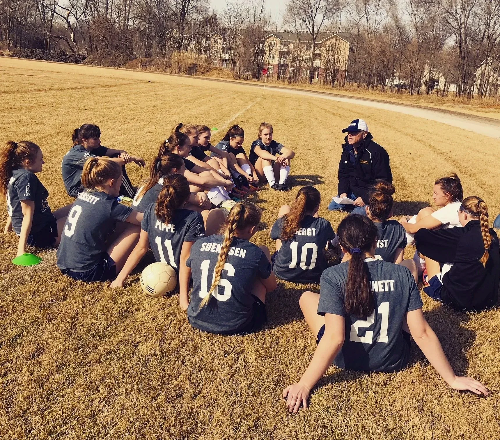 A group of female soccer players sitting on the grass listening to their coach. The players are wearing dark jerseys with white numbers, and some have soccer balls near them. The coach, wearing a jacket and cap, is speaking to them. The setting is an