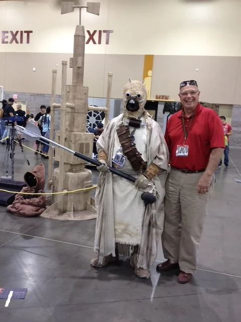 Person in a red shirt and beige pants posing with a someone dressed as a Tusken Raider from Star Wars at a convention.