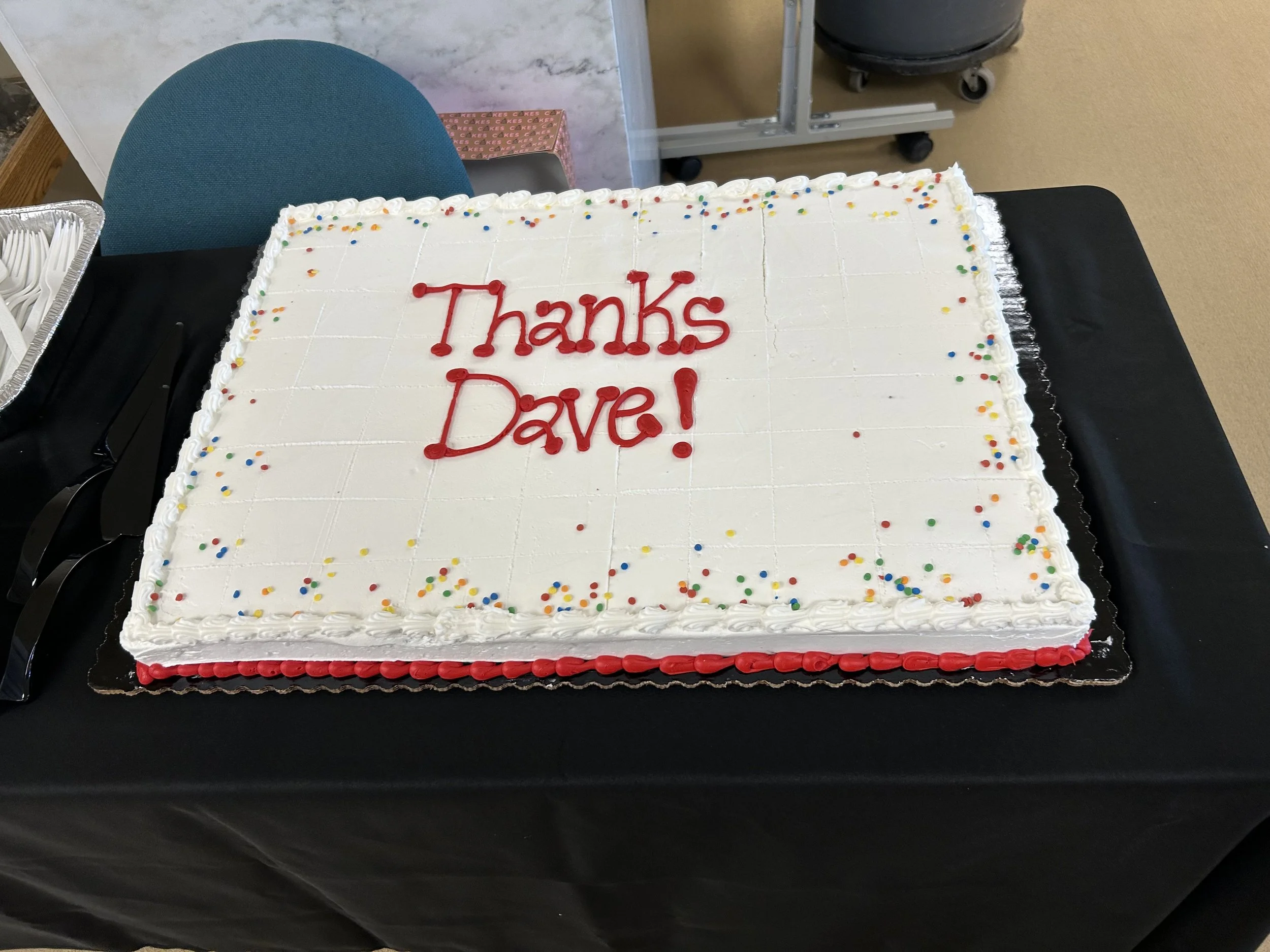 A large rectangular white frosted cake with colorful sprinkles on top and the words 'Thanks Dave!' written in red icing.