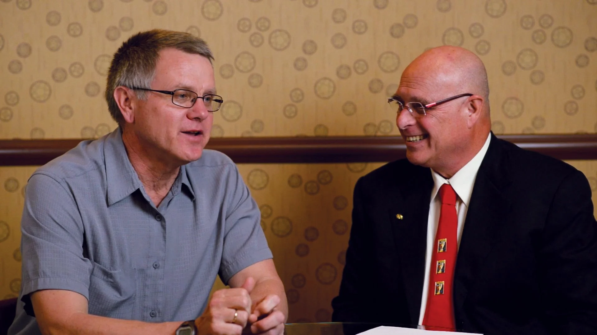 Two men sitting at a table engaged in conversation, one wearing a gray shirt and glasses, the other in a black suit with a red tie, smiling.