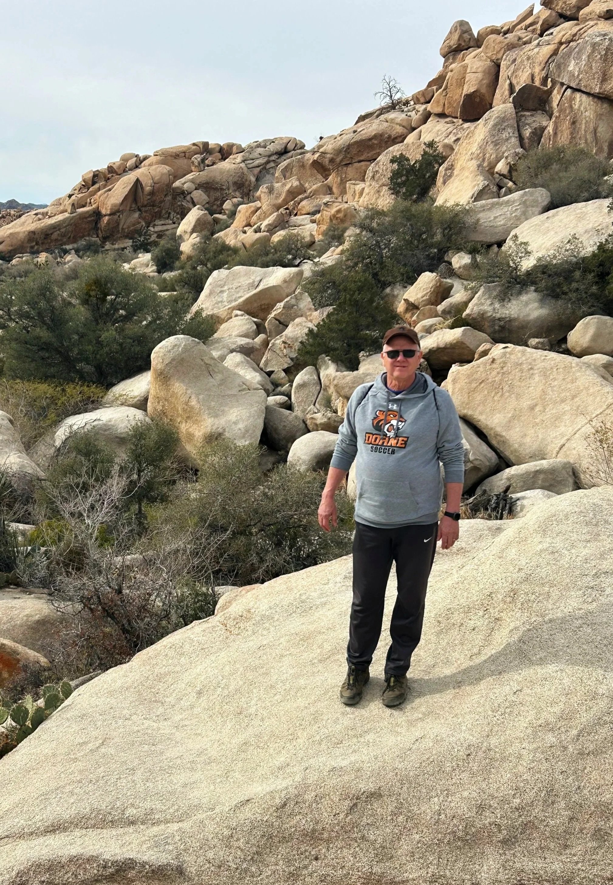 A man wearing sunglasses, a gray hoodie with a sports team logo, black pants, and hiking shoes standing on a large, smooth rock in a rocky desert landscape with boulders, desert shrubs, and sparse vegetation under an overcast sky.