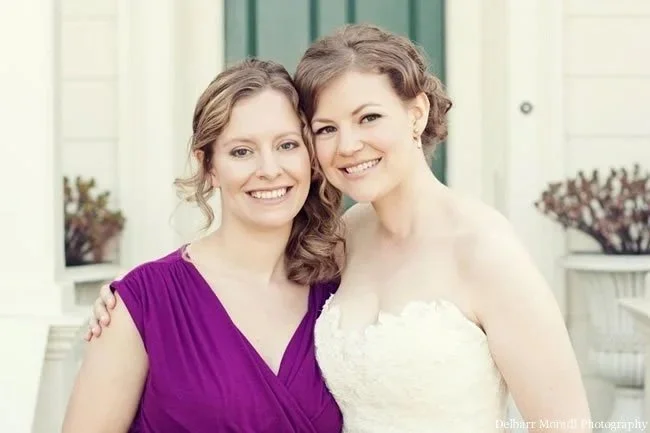 Two women smiling and embracing each other outdoors, one in a purple dress and the other in a white dress.