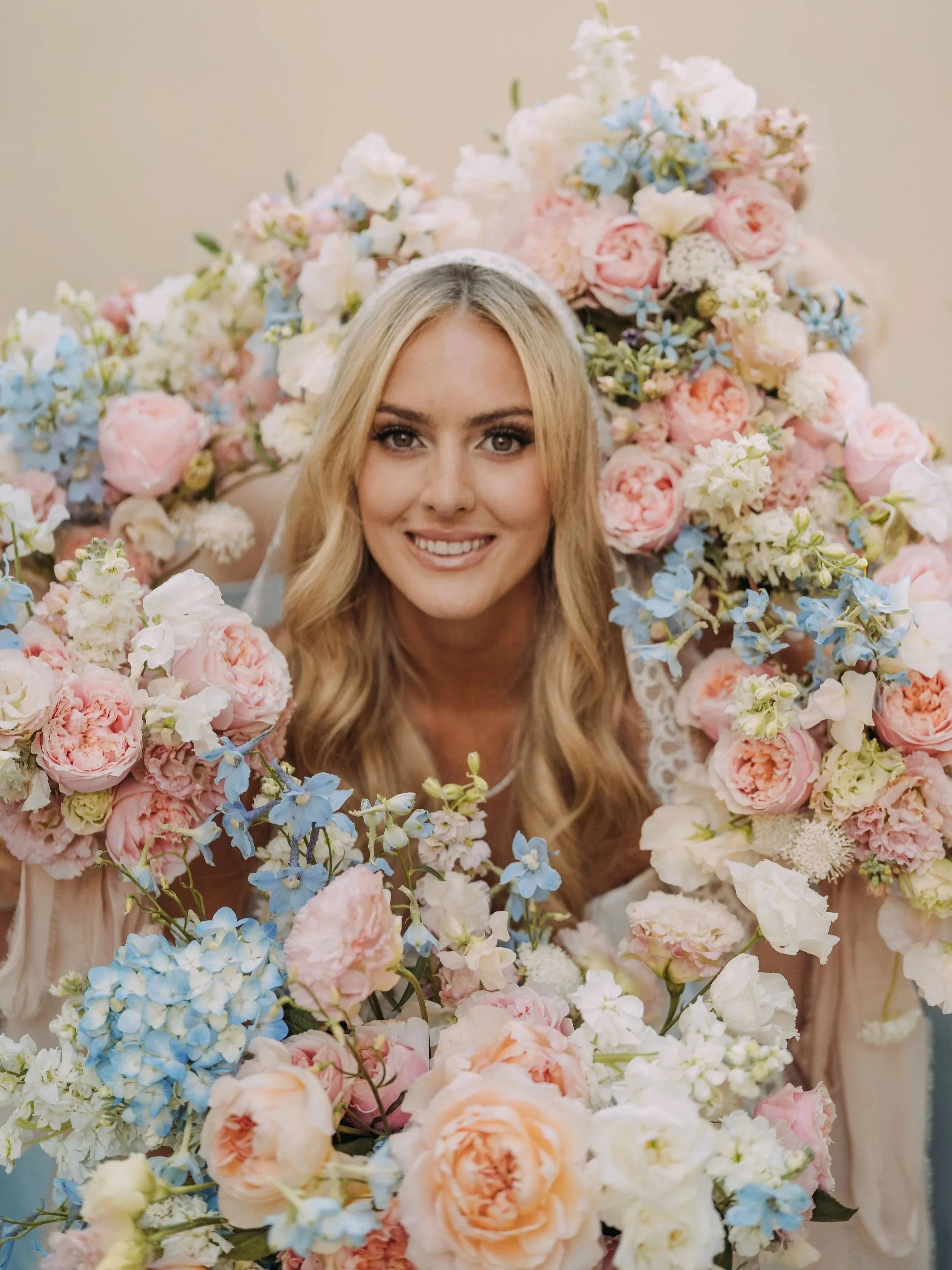 A smiling woman with blonde hair surrounded by an abundance of pink, white, and blue flowers.