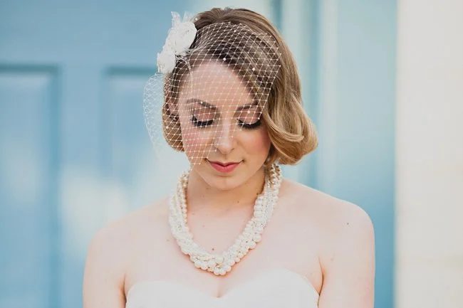 A bride with short wavy hair wearing a birdcage veil, pearl necklace, and white strapless wedding dress.