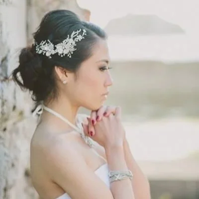 A woman with dark hair styled in an updo, wearing a floral headpiece, with her eyes closed and hands clasped in front of her face against a neutral background.