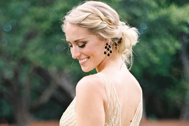 A smiling woman with blonde hair styled in an updo, wearing black earrings and a gold dress, standing outdoors with blurred green foliage in the background.