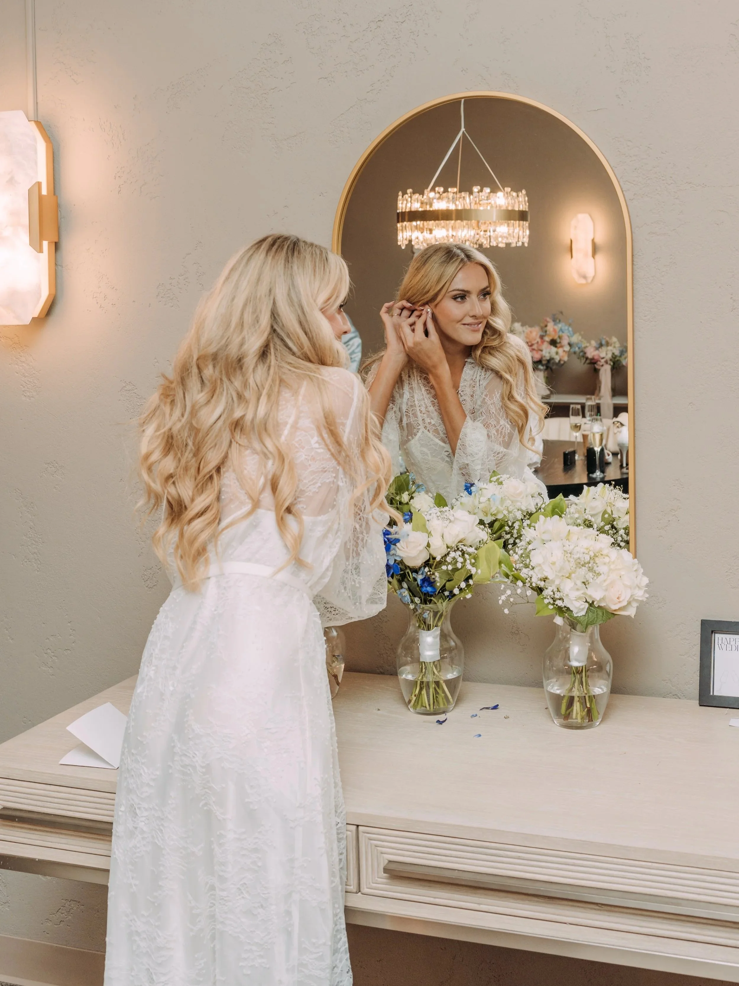 A bride on her wedding day in a white lace dress is putting on an earring while looking into a large mirror in a decorated room, with flower arrangements and vases of water on a table in front of her.