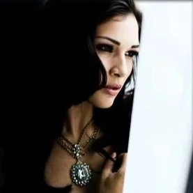 A woman with dark hair and makeup looking at a laptop screen, wearing a large pendant necklace.